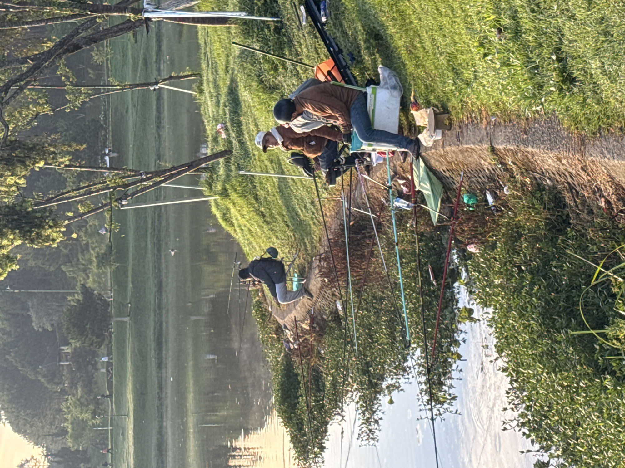 The image depicts a serene scene of several men fishing along the grassy bank of a calm lake or large pond in Da Lat, Vietnam. The warm, golden light suggests either early morning or late afternoon, creating a tranquil atmosphere with soft illumination on the foliage and water.
On the right side of the frame, a group of at least three men are seated on chairs or stools directly beside the water. They are equipped with multiple fishing rods, which extend into the water. One man in a brown jacket and blue jeans sits on a white container, while another near him wears a white cap and a brown jacket. Various fishing accessories, such as buckets, bags, and bottles, are visible on the grass around them. Further to the left, another lone fisherman, dressed in a dark jacket and grey pants, is also engaged in fishing with several rods cast.
The water is still, reflecting the sky and the surrounding trees and vegetation. The banks are covered in vibrant green grass, and dense foliage and trees line both sides of the water body. Several fishing floats are visible on the water's surface. The overall scene captures a quiet and focused activity of leisure in a natural setting. No visible text is discernible in the image.
