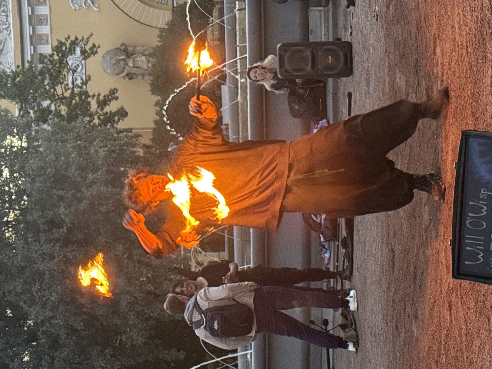 A street performer is juggling flaming objects in front of a fountain in Saint Petersburg, Russia. The performer, wearing loose-fitting dark clothing, is in the center of the frame, with flames emanating from their hands and a nearby prop. Two people are watching from the left, and another person is seated in the background to the right, smoking. The fountain, a large, tiered structure, is visible behind the performer, with water arcing upwards. The ground is a reddish-brown gravel, and trees and a building with classical architectural details form the backdrop. A sign with the words "Will O'Wisp" is visible in the foreground.
