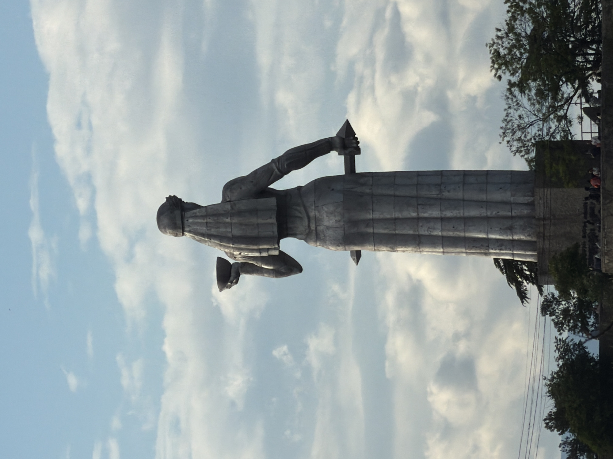 The image shows the statue of "Mother of Georgia" (Kartlis Deda) in Tbilisi, Georgia. The statue is made of aluminum and stands on a hill. It depicts a woman in Georgian national dress, holding a bowl of wine in one hand and a sword in the other. The background includes a blue sky with clouds and trees. There are people in the far background on the right, near trees, behind a concrete wall. The statue is the primary subject.