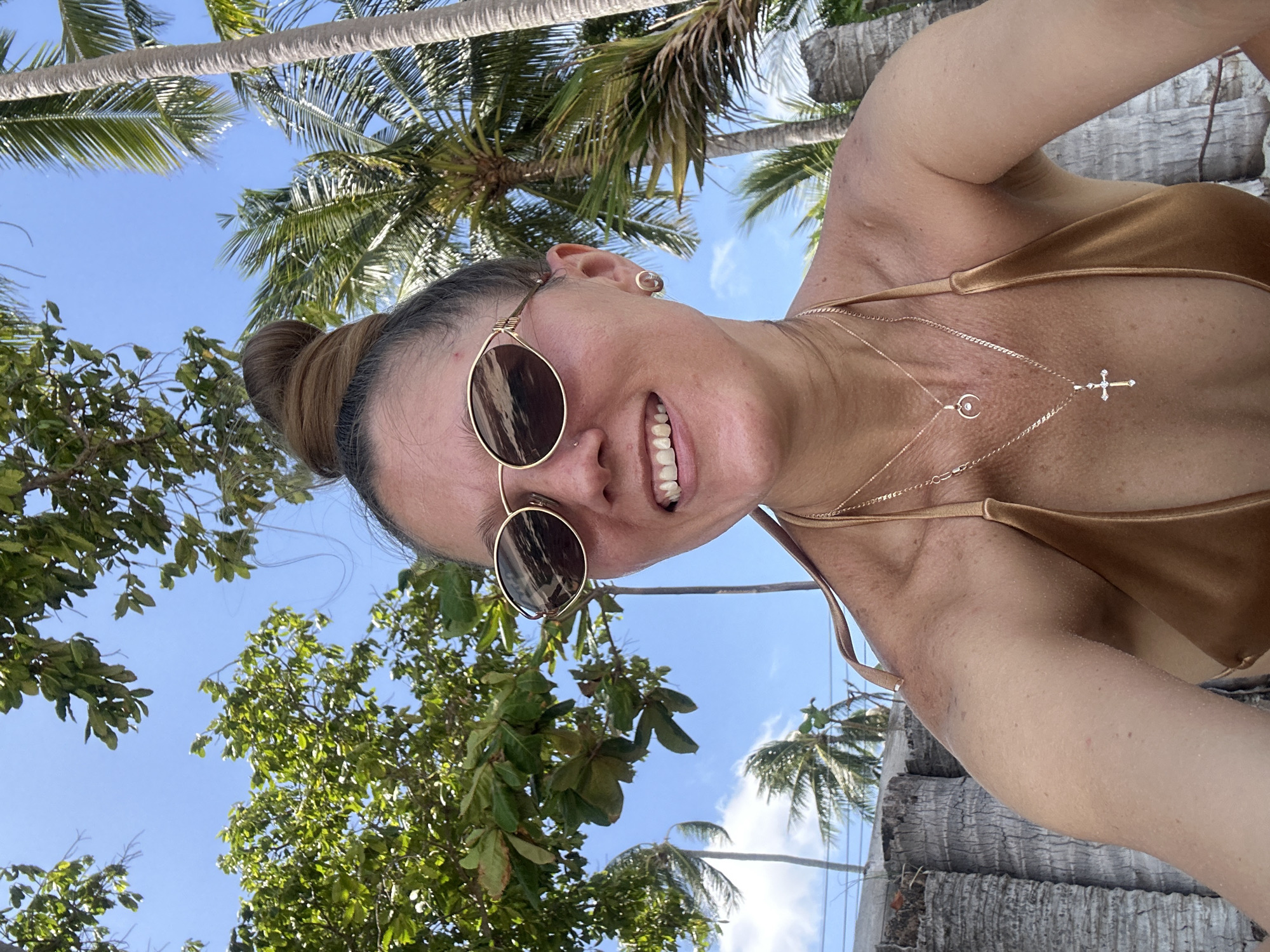 A woman with her hair in a bun and wearing sunglasses and a bikini top smiles at the camera. She is outdoors with palm trees and foliage in the background. The scene appears to be a beach.