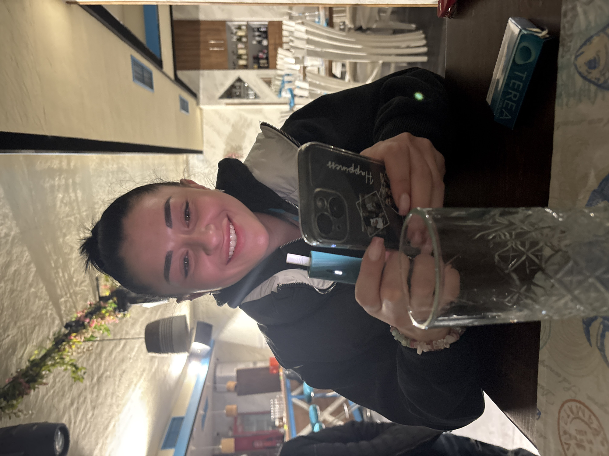 The image shows a woman holding a phone and a small electronic device in front of her. She is smiling and looking at the camera. The background shows what appears to be an indoor setting with tables, chairs, and some decorative elements. On the table next to her is a blue box of Tereas. The location in the city is Unknown, Unknown.