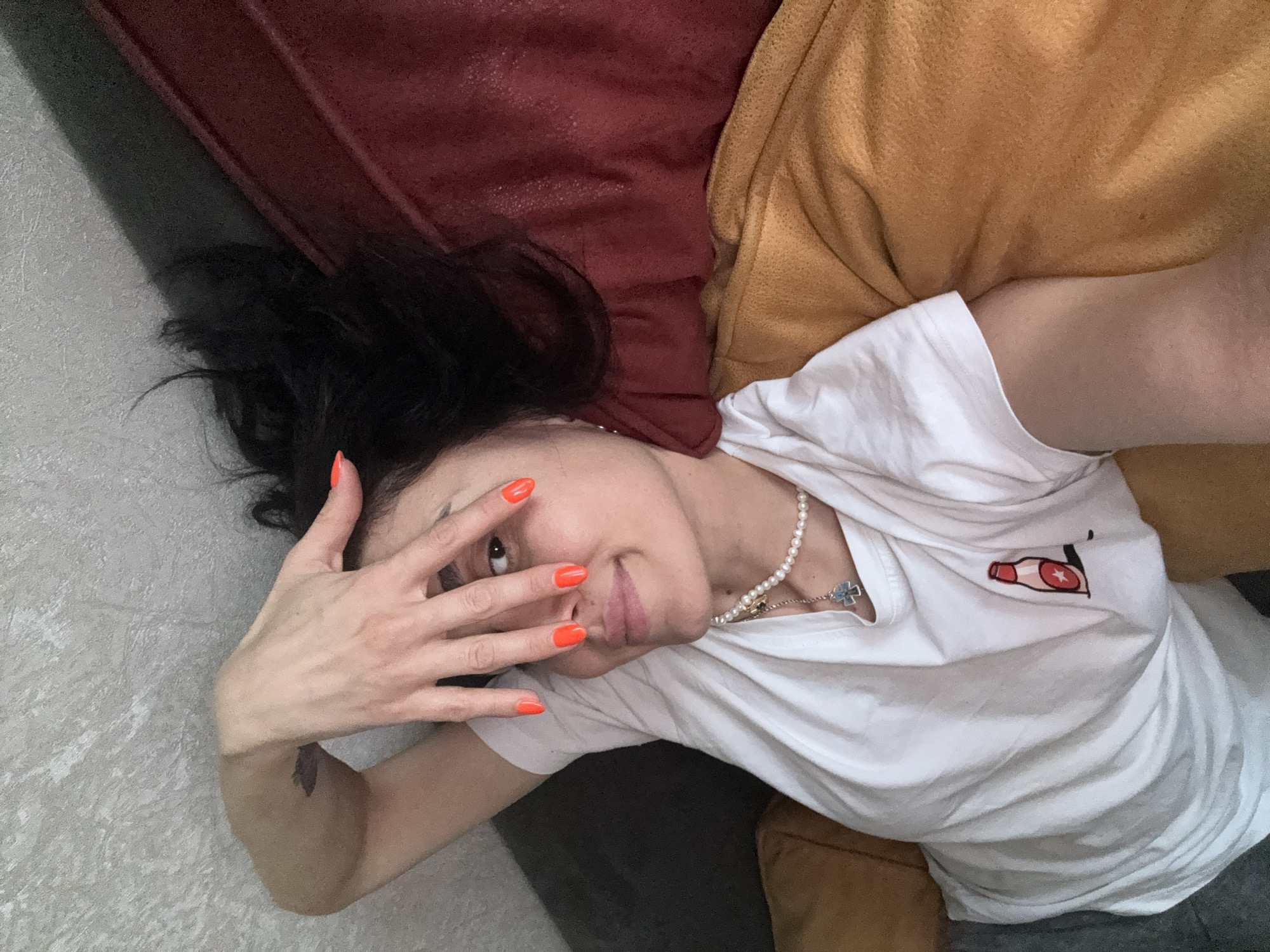 The image shows a young woman lying on a couch. She has dark hair and is wearing a white t-shirt with a small graphic of a bottle on it, and a pearl necklace. Her right hand is covering part of her face, revealing an orange nail polish. The couch appears to be a dark red or maroon color, with a mustard yellow blanket draped over it. The background is a textured gray wall. The image was taken in Tbilisi, Georgia, in an indoor setting.