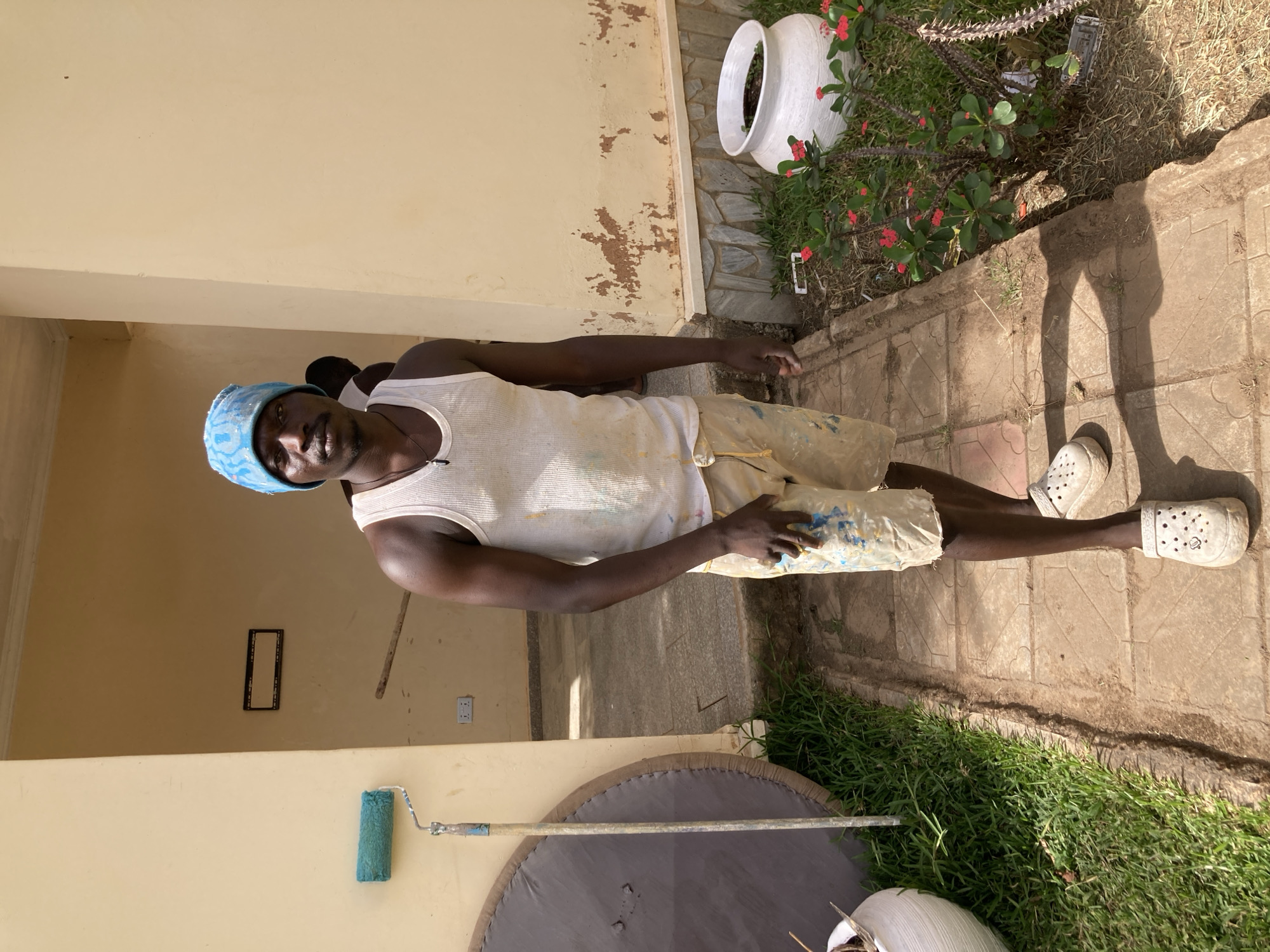 A man stands outdoors in a bright, sunny setting, looking directly at the camera. He is dark-skinned and wears a light blue patterned beanie, a white ribbed tank top heavily splattered with paint, and paint-stained khaki shorts. His feet are clad in white Crocs. He holds a plastic bag or container, also marked with paint, in his right hand.
The scene appears to be an outdoor area, possibly a courtyard or veranda in Jos, Nigeria. The building walls are a light yellow or cream color, with some areas showing peeling paint and a darker underlying layer. The ground is paved with light brown interlocking tiles, bordered by a strip of grass and dry foliage. In the foreground, a blue-green paint roller with a long handle leans against a wall, near a large, round, grey mat on the ground. In the background, a white ceramic pot or planter sits amidst a thorny shrub bearing small red flowers. A small rectangular frame and an electrical outlet are visible on the wall behind the man.
The strong, distinct shadows cast by the man and objects indicate bright daylight, likely late morning or early afternoon. The man’s attire and the presence of painting tools suggest he is engaged in painting activities. His expression is calm and neutral. No visible text is present in the image.