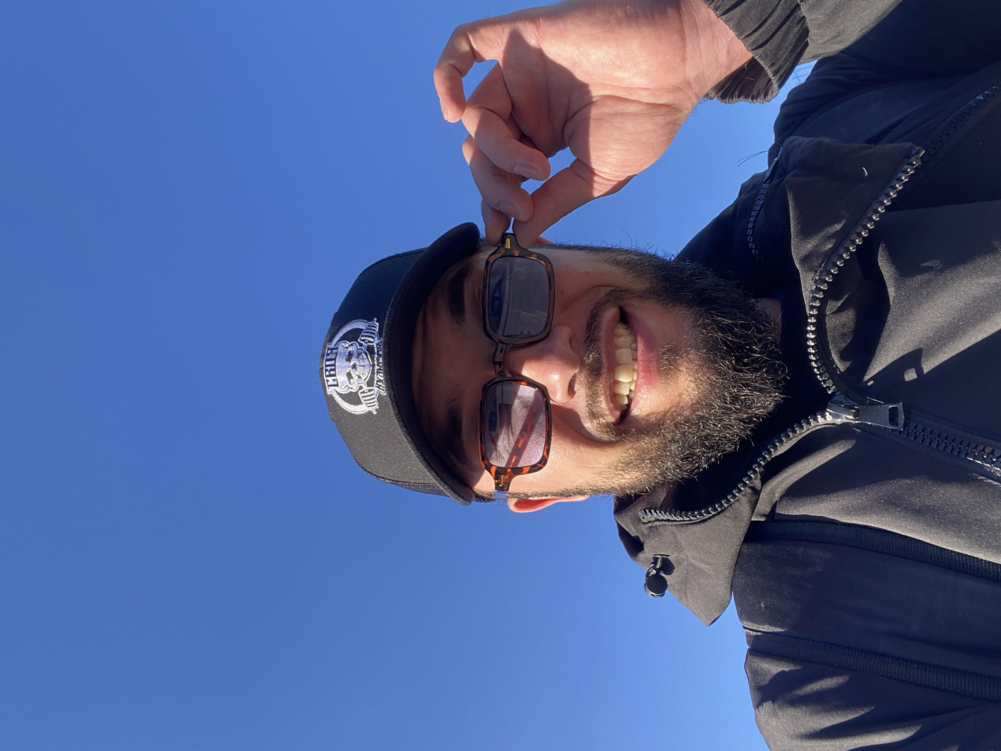 A man is photographed outdoors against a clear blue sky. He is wearing a black baseball cap with a white logo, brown-rimmed rectangular sunglasses, and a black jacket. The man has a beard and is smiling, with his right hand adjusting his sunglasses. He is looking directly at the camera. The location is Koskolovo, Russia.