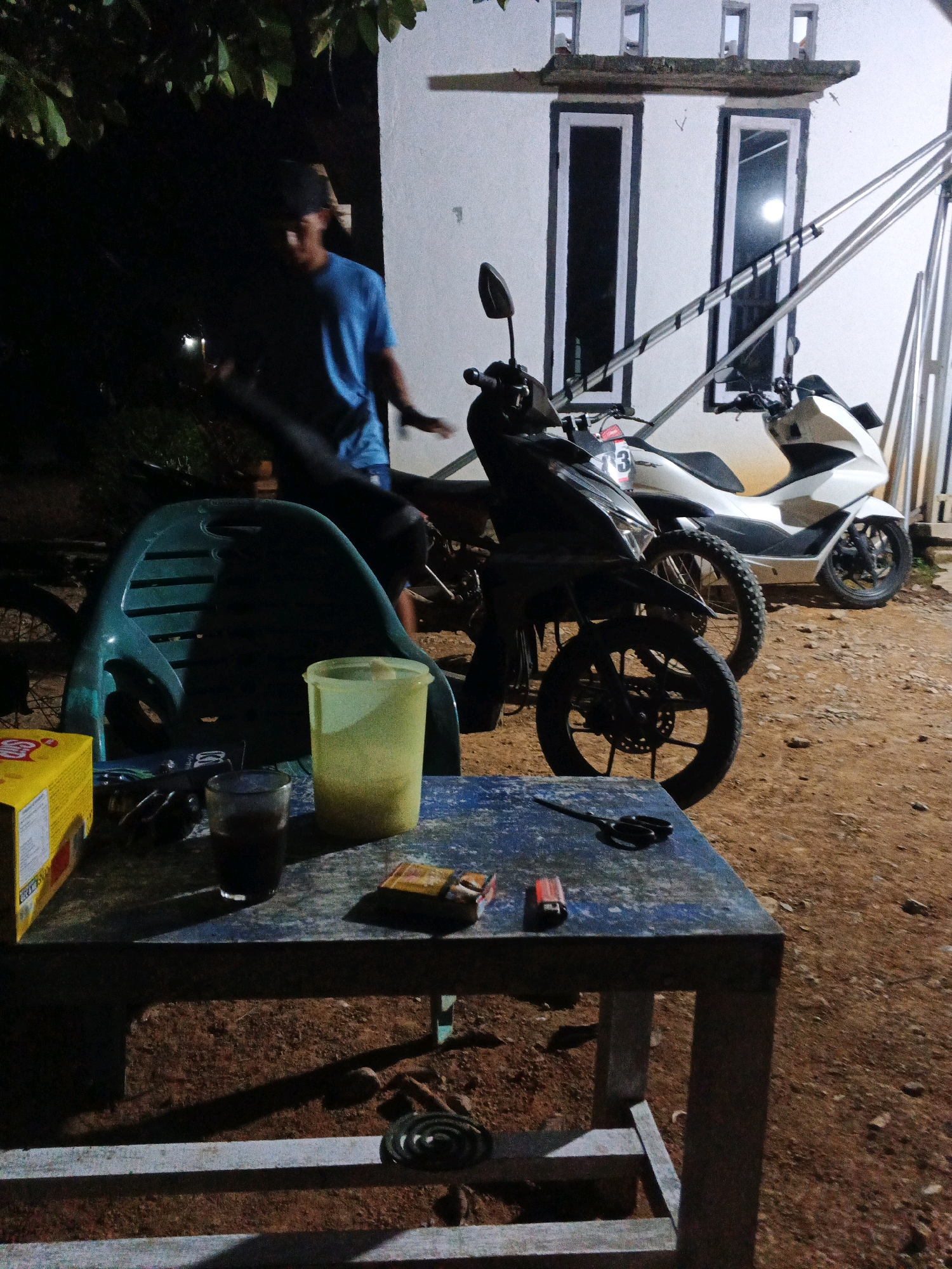 In the dimly lit outdoor setting of Brangan Mulya, Indonesia, a person wearing a blue t-shirt and a dark cap is visible standing near a white building. In the foreground, a weathered blue table displays several items: a glass of dark liquid, a yellow box, a pack of cigarettes, a lighter, and a pair of scissors. Behind the table, a green plastic chair is positioned. Two motorcycles, one black and one white, are parked near the building. The ground is covered with dirt and gravel. The scene appears to be captured at night.