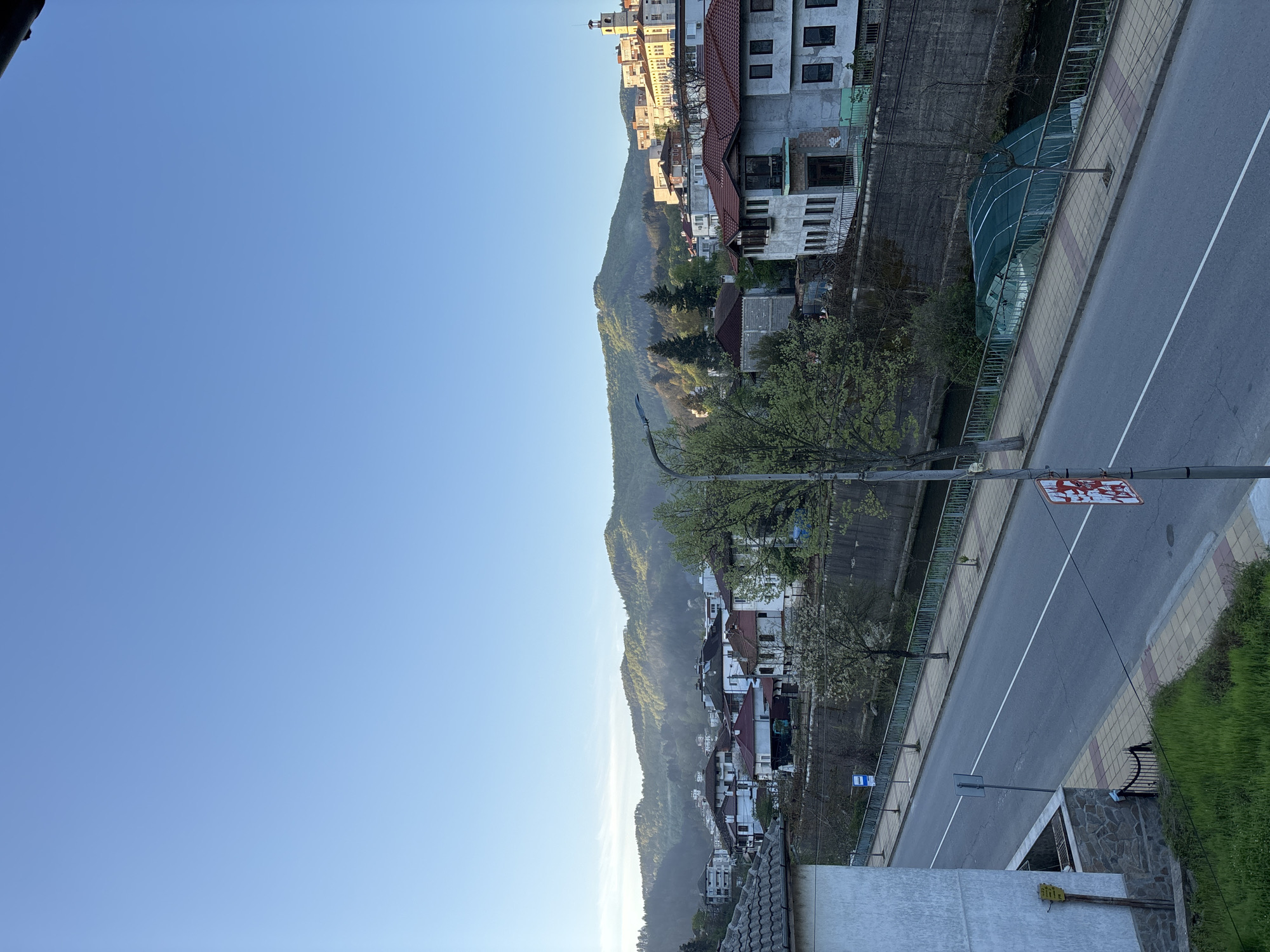The image captures a scenic view of Zlatograd, Bulgaria, with a town nestled at the base of a verdant mountain range under a clear blue sky.
In the foreground, a paved road with a distinct white center line extends from the bottom right towards the upper left. To the right of the road is a sidewalk paved with yellow and reddish tiles, bordered by a metal fence. A grassy area and a stone wall are visible further to the right. A street light pole stands prominently on the right side of the road.
On the left side of the road, another metal fence separates it from what appears to be a narrow river or canal, beyond which various buildings are constructed. These houses typically feature white facades and red or brown tiled roofs, climbing up the lower slopes of the mountain. Green trees, some with fresh spring foliage or blossoms, are interspersed among the buildings and along the waterway. A large, dark green netted structure, possibly for agriculture or construction, is visible near the water on the left.
The mid-ground and background are dominated by a massive, forested mountain, densely covered in green trees, which stretches across the horizon. The top of the mountain meets a vast, uninterrupted expanse of bright blue sky, indicating clear weather.
The scene appears peaceful and devoid of human activity. The soft, natural light suggests either early morning or late afternoon, creating a gentle illumination over the landscape.
Visible text includes a rectangular red and white sign on a pole by the roadside, which appears to be a traffic or informational sign, but the white text or symbols against the red background are indistinct. Further down the road on the left, a small blue and white sign, possibly indicating a bus stop, is also visible.