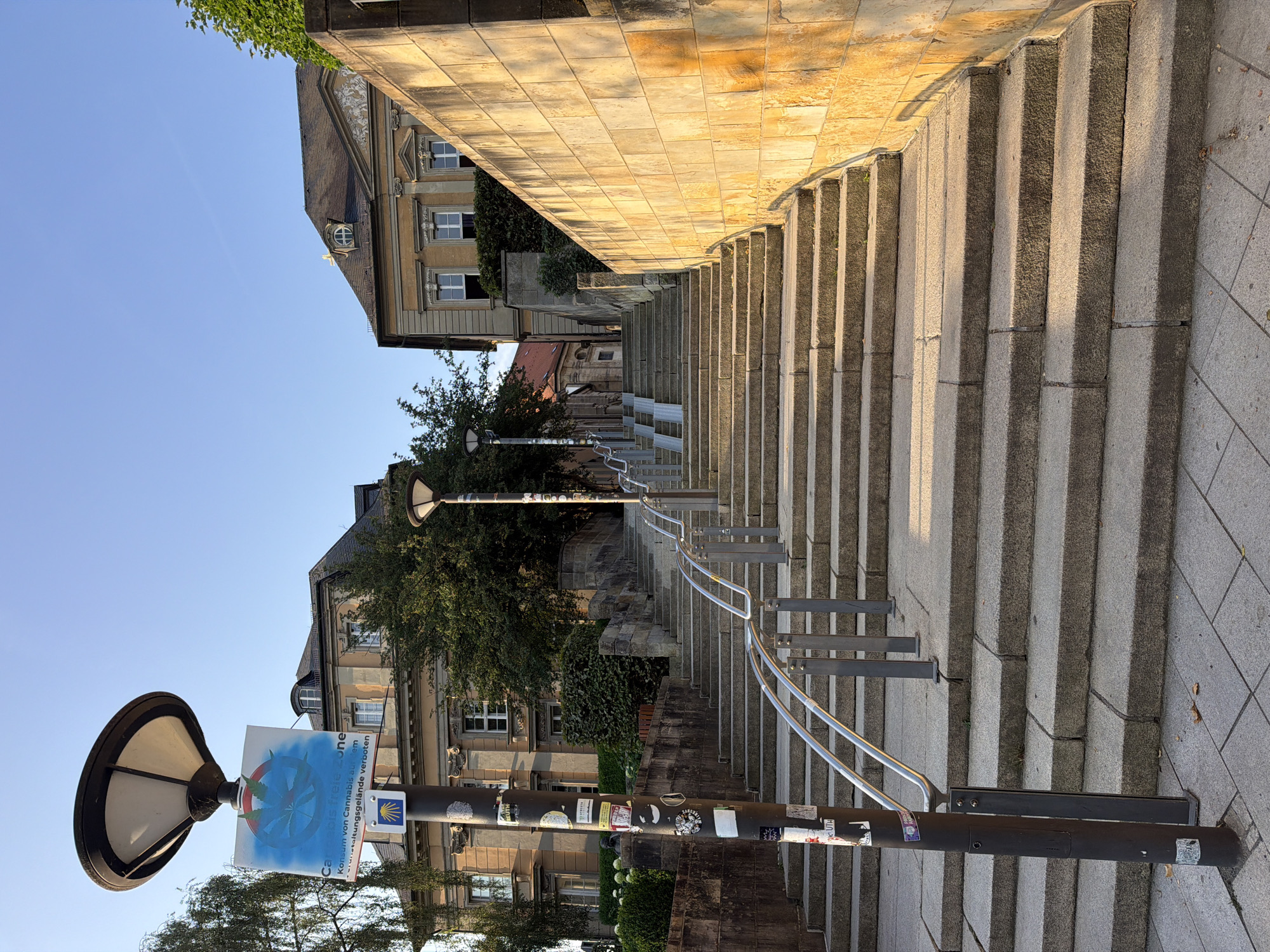 The image shows a wide, stone staircase leading upwards. A black lamppost with a sign reading "Cannabis Freie Zone" is on the left. The staircase is bordered by a stone wall on the right, illuminated by sunlight. In the background, there are buildings with traditional architecture, one appearing to be a historical building with ornate detailing. Trees and vegetation are visible, and the sky is clear and blue. The scene appears to be in a public area, possibly a park or a historical district. Based on the sign mentioning "Cannabis Freie Zone" and German text, the location is likely Germany. The presence of the sign and the architectural style suggest a European city, and specifically, the prompt identifies the location as Bayreuth, Germany. There are no people visible in the image.