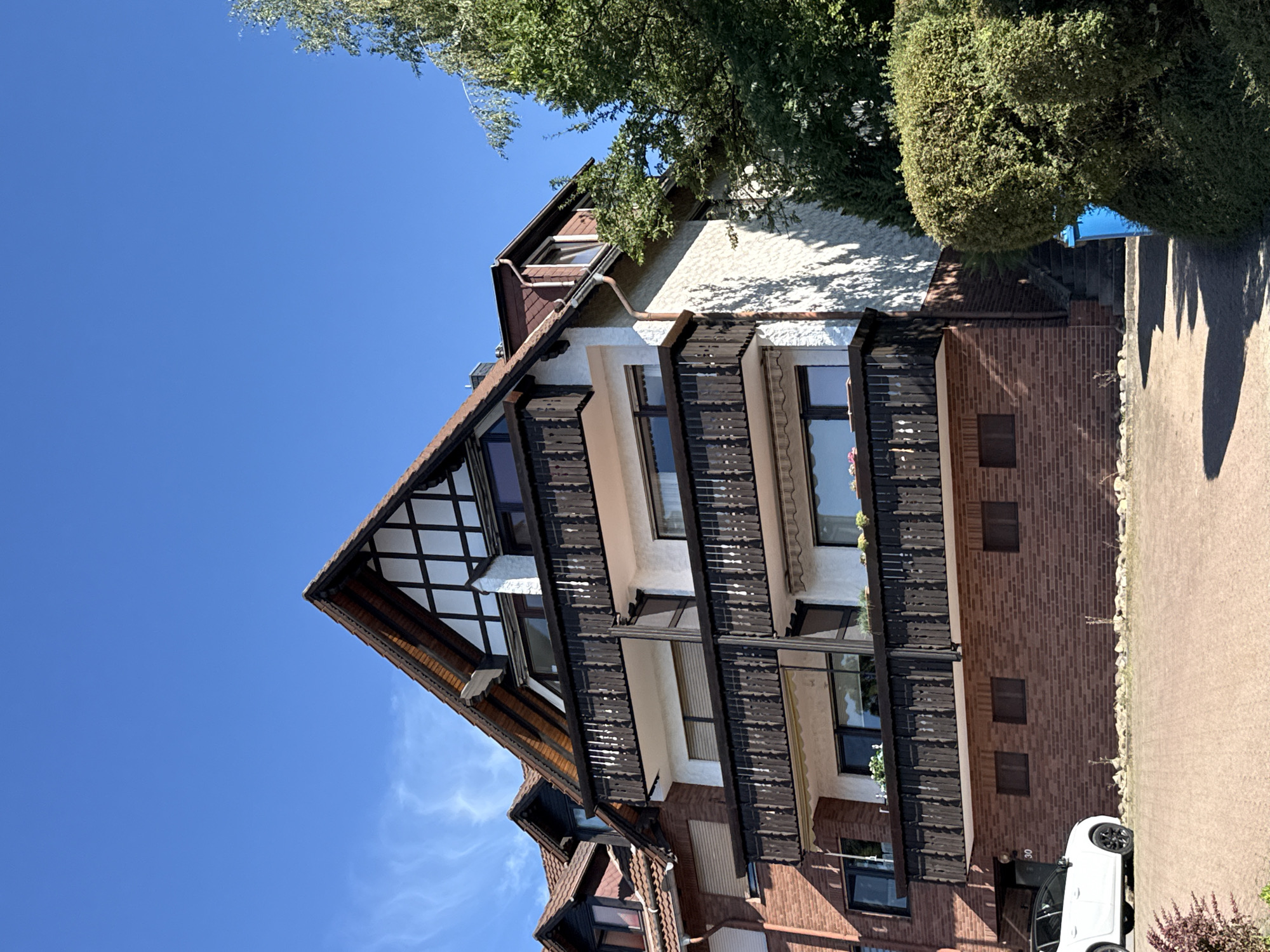 A multi-story building with a tiled roof and balconies is depicted under a clear blue sky. The building features a mixture of brick and white stucco facades. The gabled roof has exposed wooden beams, a common architectural style. There are several balconies with dark wooden railings, some of which have awnings. A white car is parked in the foreground on a paved driveway. A bright blue recycling bin is visible next to a large, well-maintained green hedge. Stairs leading up the side of the building are partially obscured by the hedge. The scene is set in the town of Bad Sachsa, Germany.