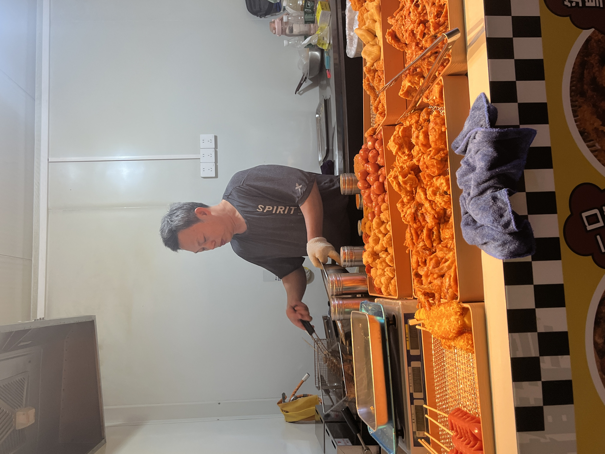 A person is preparing fried foods at a food stall. The person is wearing a grey t-shirt and white gloves, using tongs to handle food in a deep fryer. Assorted fried items are displayed in bins, including fried chicken, sausages, and other snacks. A blue towel is nearby. The stall has a black and white checkered design. The location is Jiangjin, China.