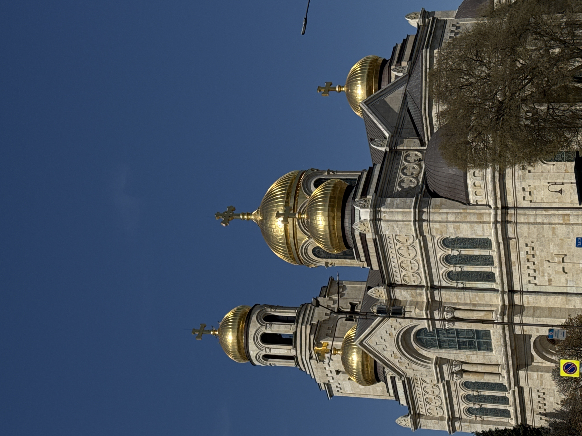 An outdoor, bright daylight shot depicts a grand Orthodox church in Varna, Bulgaria, set against a clear, deep blue sky. The majestic building is constructed from light-colored stone, featuring elaborate architectural details including arches, columns, and decorative patterns on its facade. It boasts multiple golden, ribbed onion domes, each crowned with a golden cross. Two prominent towers, each topped with a golden dome and cross, frame the central structure. Numerous windows with dark grilles are visible along the building's sides.
The photograph is taken from a low angle, emphasizing the church's height and intricate design. On the right side of the building, a deciduous tree with bare branches partially extends into the frame, suggesting autumn, winter, or early spring. A utility pole or street light fixture is faintly visible in the upper left portion of the sky.
Visible text includes a blue street sign on the building's upper right facade with the word "ТАКСИ" (TAXI) written in Cyrillic. Below this, a yellow square sign with a blue circle and a red diagonal line, indicating "no stopping" or "no waiting," is present near the bottom right. The bright sunlight suggests midday or afternoon, casting clear shadows on the detailed stonework.