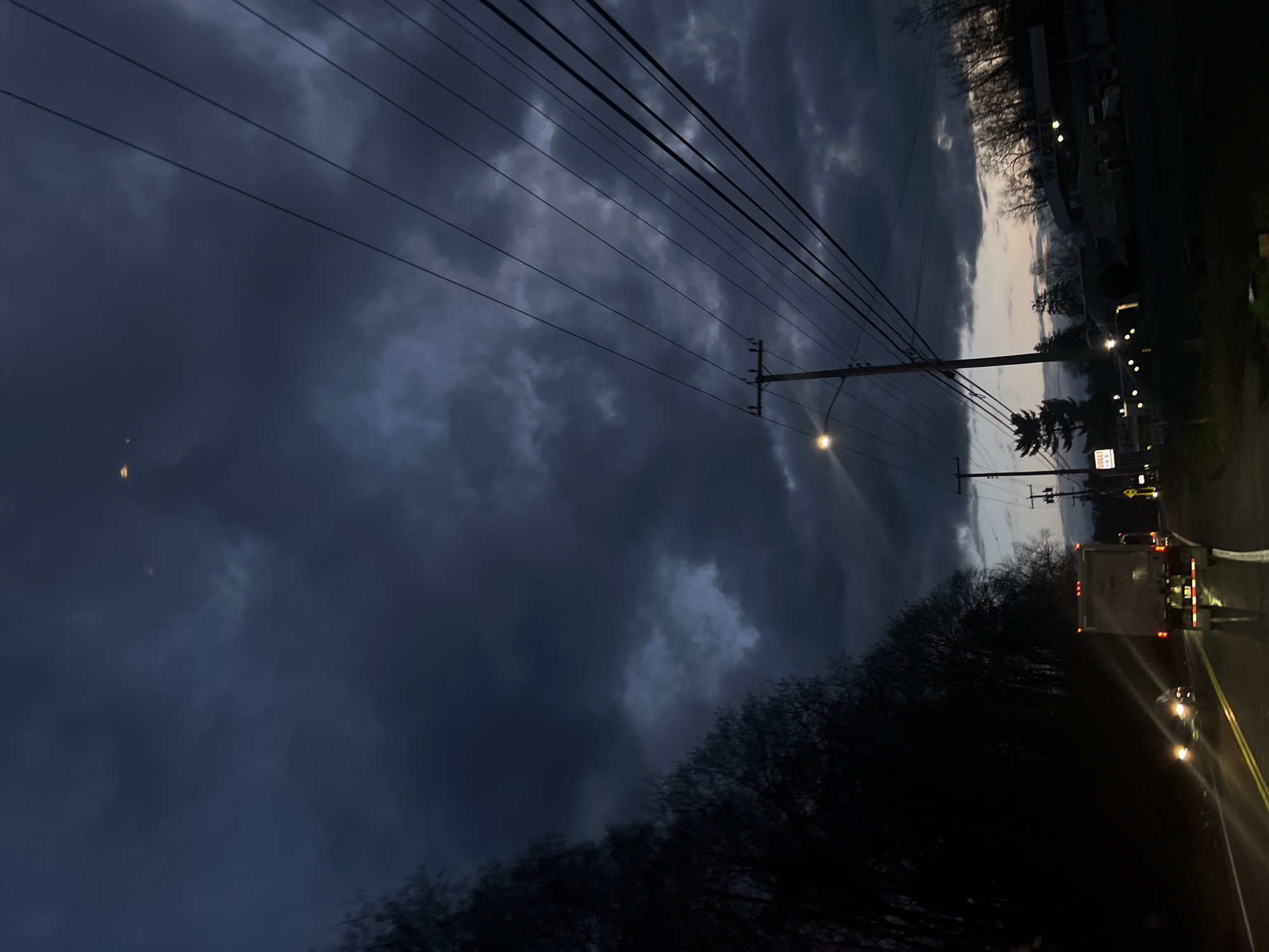 The image captures a scene at dusk on a road in Balmville, United States of America. Overcast skies are filled with dark, dramatic clouds. Overhead power lines stretch across the scene. A streetlight illuminates a pole. A white truck with rear lights is on the road along with other cars, with headlights visible. Bare trees line the side of the road. Signs and buildings are visible in the background.