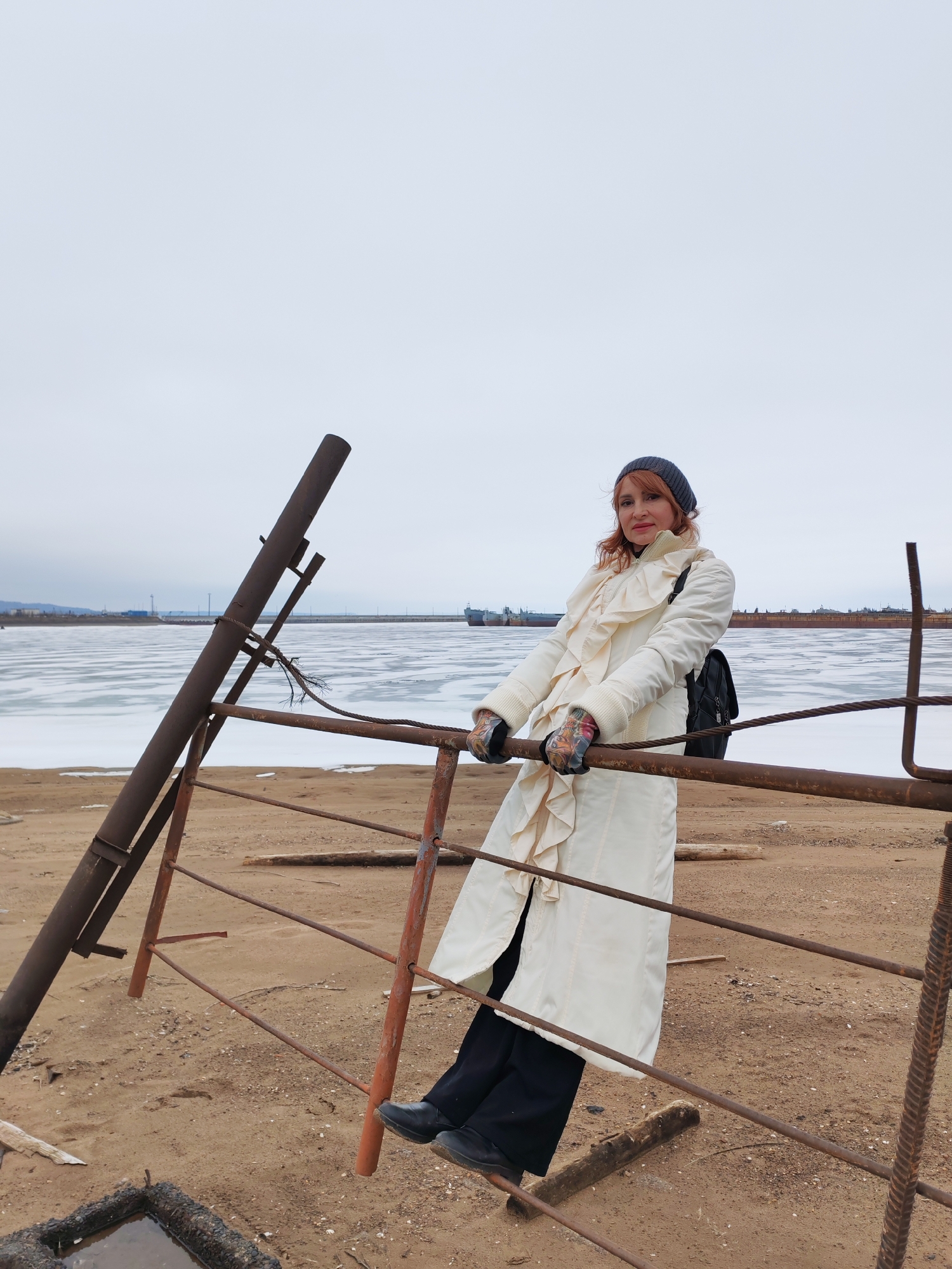 A woman is standing on a beach in Chaikovsky, Russia, leaning on a rusty metal structure. The woman is wearing a white coat with ruffles, black pants, black shoes, a grey knitted hat, and gloves with a floral pattern. She has reddish-brown hair. Behind the woman is a frozen body of water with several ships visible in the distance. The sky is overcast. The beach is sandy and littered with debris. The metal structure appears to be part of an old dock or pier.