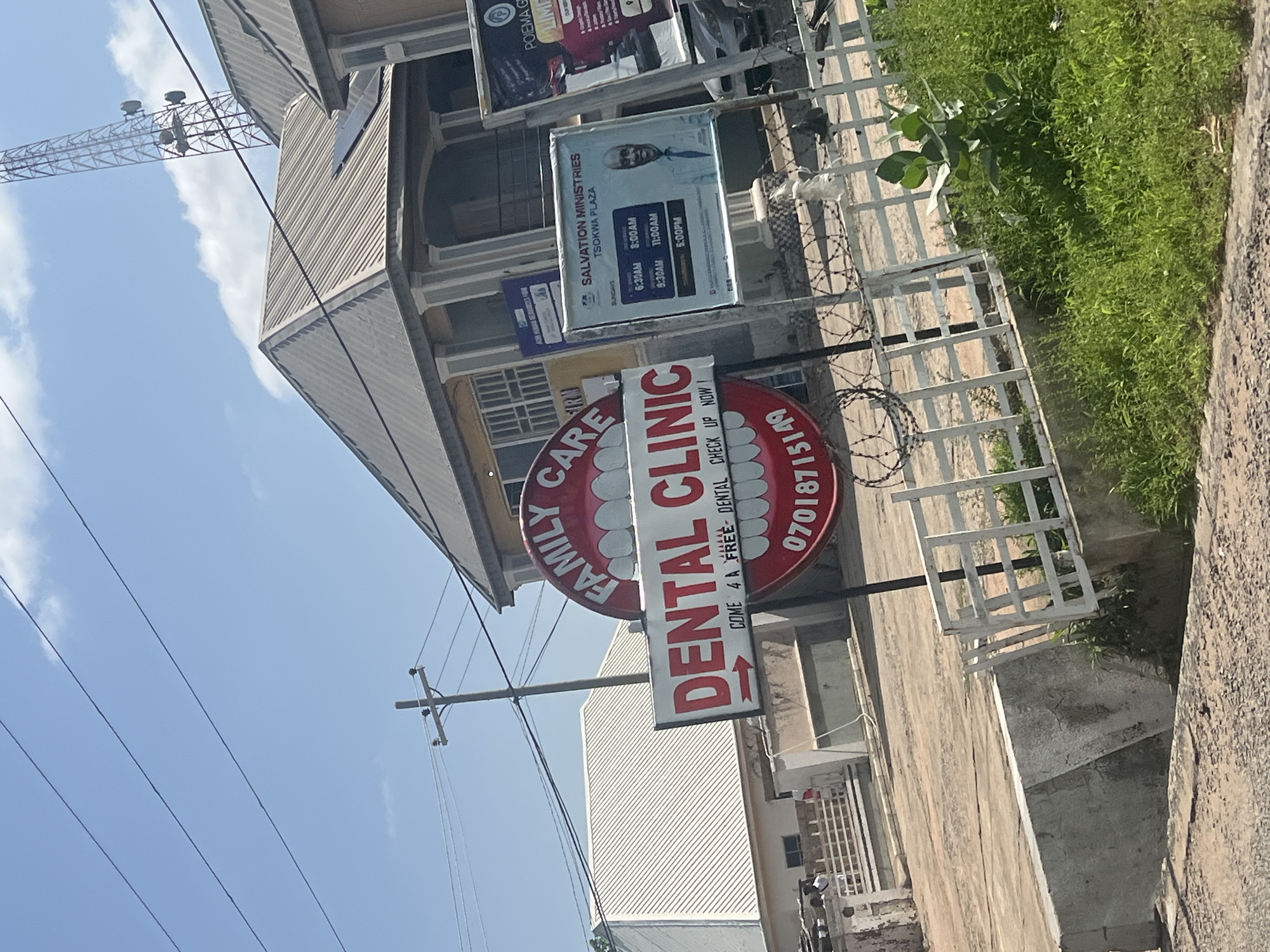 The image captures a street scene in Jalingo, Nigeria, focusing on a dental clinic and a nearby church advertisement. A large, circular red and white sign for "FAMILY CARE DENTAL CLINIC" dominates the foreground, with text in red stating "COME 4 A FREE DENTAL CHECK UP NOW!" and a phone number below. Adjacent to the dental clinic sign is a smaller, rectangular sign for "SALVATION MINISTRIES TSOKWA PLAZA," displaying service times and a picture of a man. Further to the right, another advertisement is partially visible. The background features buildings with corrugated metal roofs and a cell phone tower in the distance. Overhead power lines crisscross the sky, and the scene is set against a clear blue sky with some clouds. A white fence with barbed wire separates the foreground from the background. The environment appears to be an urban or semi-urban area, with evidence of infrastructure like utility poles and power lines. The overall impression is one of a typical commercial and religious area in Nigeria.