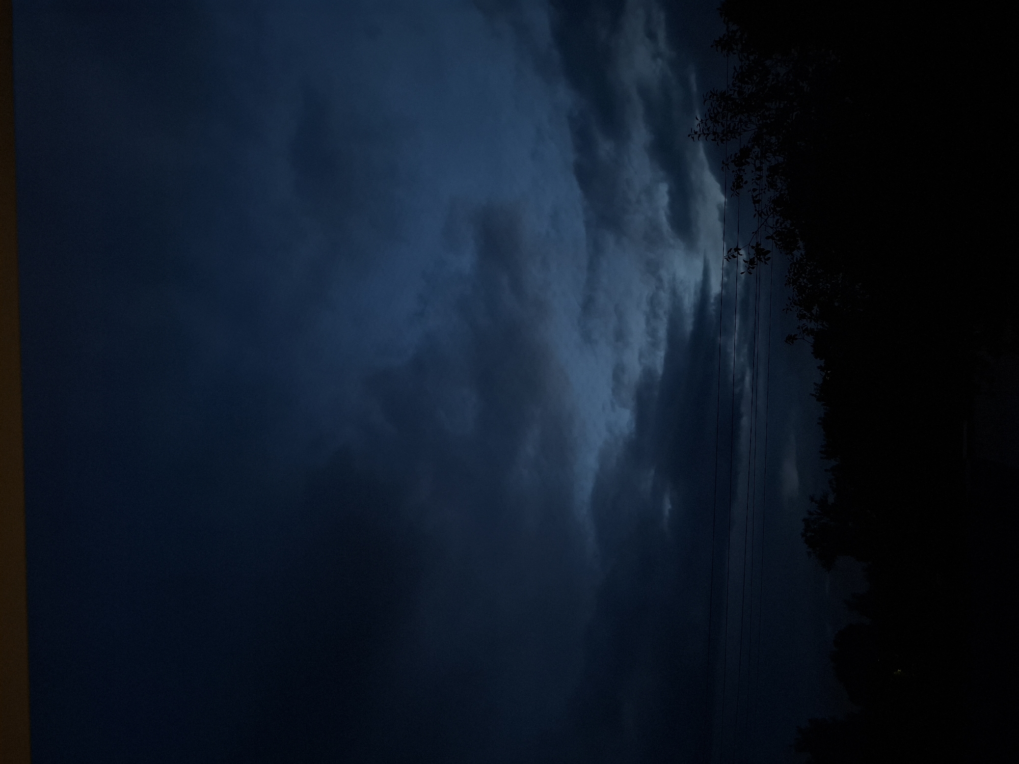 The image captures a dark, cloudy sky with muted blue and gray tones. In the distance, a dark silhouette of trees and foliage forms the horizon line. Thin, dark horizontal lines, possibly power lines, are visible against the sky in the mid-ground. The overall atmosphere is dim and somber, suggesting either dusk or a heavily overcast day. The location is identified as Novlyanka, Russia.