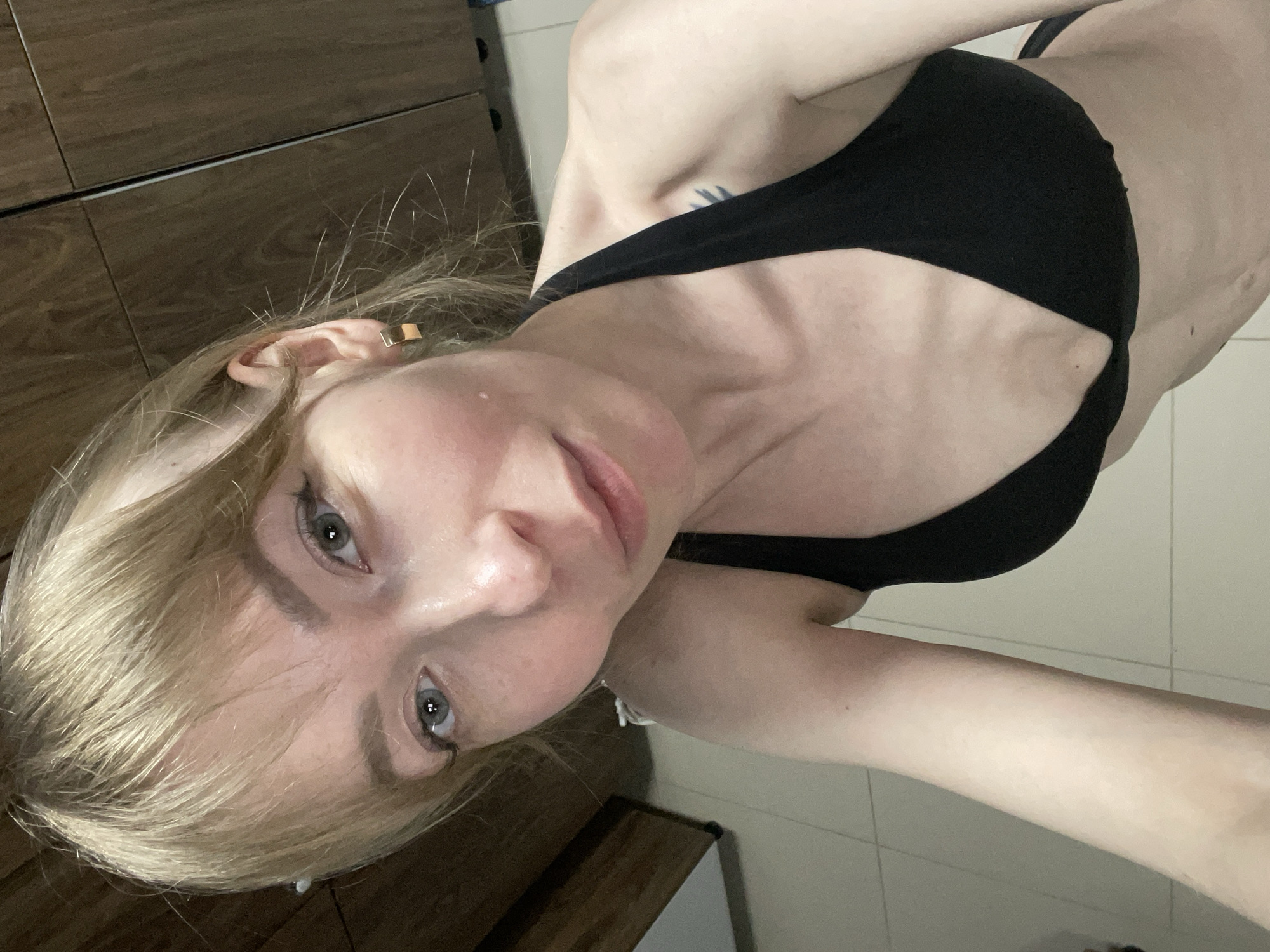 The image is a selfie taken by a woman with long, light blonde hair and light skin. Her head is tilted, and she looks directly into the camera with light blue-grey eyes and a neutral expression. She is wearing a black bra or bikini top. A gold-colored ear cuff is visible on her left ear, nestled within her hair.
A dark, linear tattoo is visible on her upper right shoulder or back area, partially obscured by her arm and the black top. The pose suggests she is angling her body to capture herself from the chest up, with part of her arm and torso visible.
The background indicates an indoor setting, possibly a bedroom or bathroom. On the upper left, there is dark wooden furniture, appearing to be a dresser or wardrobe. The lower and right portions of the background show light-colored tiled surfaces, either a wall or a floor. There is no visible text in the image. The scene is located in Protasy, Russia.