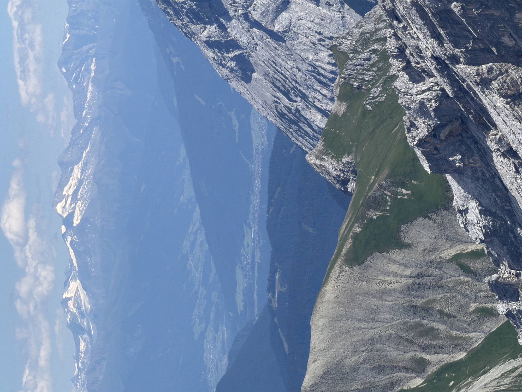 The image shows a panoramic view of a mountain range. In the foreground, there are steep, rocky slopes with patches of green grass. The middle ground features rolling hills covered in dark green forests, with a small village nestled in a valley. The background reveals a majestic mountain range with snow-capped peaks under a bright blue sky with scattered clouds.
While the image depicts a mountain landscape, it is not possible to definitively pinpoint the exact location within Garmisch-Partenkirchen, Germany, or any specific city, without additional information. The scene appears to be a natural mountain vista. There are no people or specific objects that would indicate a particular urban location.