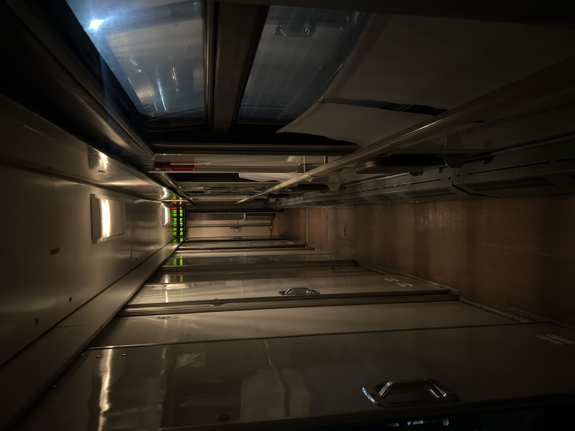 The image shows the interior of a train compartment. The view is looking down a narrow corridor with multiple compartments visible on both sides. The compartments have overhead luggage storage. The walls are light-colored. There are windows or skylights in the ceiling. The compartments appear dark. There are some small lights visible along the corridor. There is no evidence to confirm the location.