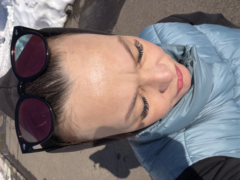 An overhead photograph captures an adult female outdoors in Chaikovsky, Russia. The individual is positioned looking downwards, her eyes closed or heavily downcast, accentuated by prominent false eyelashes. Her eyebrows are dark and her brow shows a slight furrow. Her lips are closed and adorned with reddish-pink lipstick. Resting on top of her head are black-framed sunglasses with reflective, reddish-hued lenses, which exhibit visible cracks or scratches. She wears a dark hooded garment pulled back on her head, a light blue quilted puffer vest, and a dark long-sleeved shirt underneath. The immediate environment consists of a dark, paved surface, likely asphalt, with patches of melting snow visible to the right and in the background. Direct sunlight casts a distinct shadow of the individual onto the ground to the left.