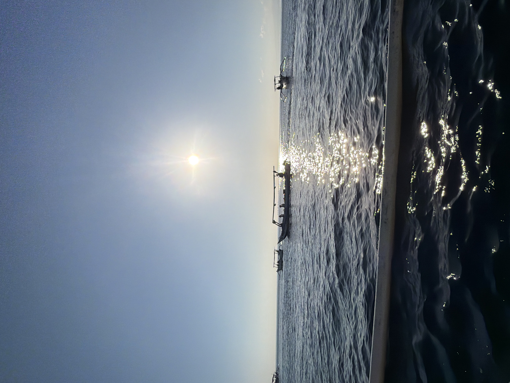 The image depicts a serene seascape at dawn or dusk, with the sun positioned high in the clear blue sky, casting a bright glare. The sun's reflection shimmers on the gently rippling, dark blue water. Several traditional outrigger boats, likely located off the coast of Seririt, Indonesia, are visible on the water. Some boats are empty, while others appear to carry passengers, suggesting early morning or late afternoon fishing or transport activities. The foreground shows a portion of another boat, indicating the photograph was taken from the water. The horizon is a faint line where the water meets the sky.