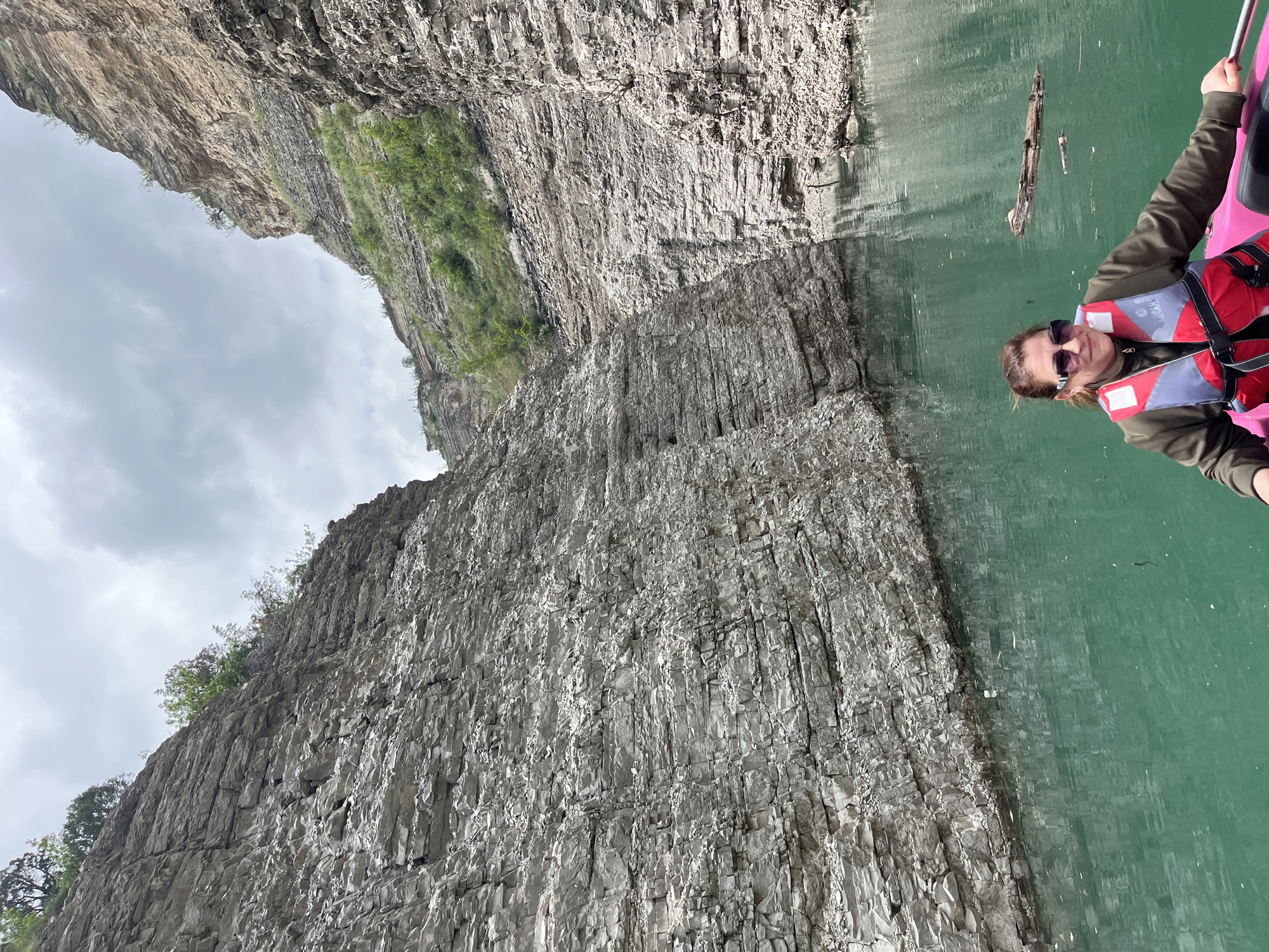 The image shows a person wearing sunglasses and a life vest, possibly in a kayak or small boat, on a body of water with tall, rocky cliffs alongside. The water appears greenish and there's some debris floating on the surface. The cliffs are gray and layered with visible rock formations. There are some patches of vegetation on the cliffs. The sky is overcast. There is no visual information within the image to suggest the location is in the city of Druzhba, Russia.