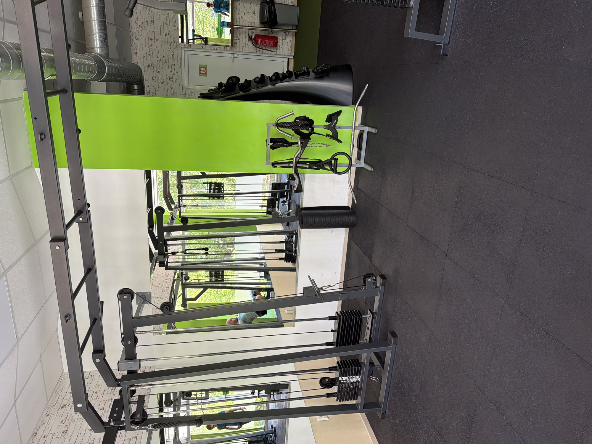 The image captures the interior of a gym. The floor is covered with black rubber mats. Prominent gym equipment includes a cable crossover machine with weight stacks and various cable attachments. The walls feature a combination of white brick and light green paint. A mirror reflects the gym interior, including individuals exercising. Above, there's visible ductwork and near the ceiling there is a red fire extinguisher. Additional equipment is positioned against the wall. The location is a gym in the city of Sofia, Bulgaria.