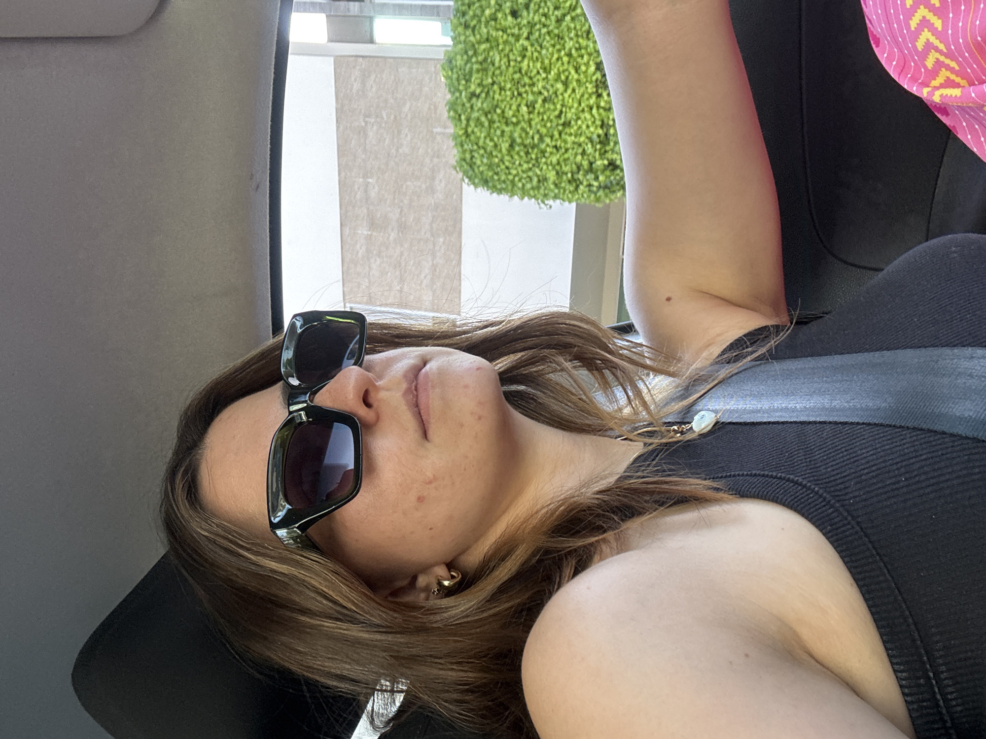 A medium close-up, slightly tilted portrait shows a person reclining in what appears to be the backseat of a vehicle during the daytime. The person, with light brown hair, is wearing dark, rectangular sunglasses and a black ribbed tank top. A seatbelt is visible across their chest and shoulder. A small gold hoop earring with a star charm is visible on their right ear. Their face is turned slightly upwards, with their lips slightly parted. Small skin imperfections are visible on their cheek and chin area.
The background inside the vehicle is largely out of focus, showing a grey or light-colored interior trim on the left. Through the vehicle's window on the right, a bright, sunny outdoor scene is visible. There's a neatly trimmed, vibrant green shrub or topiary, suggesting a well-maintained outdoor area like a park or street. A tall, light-colored building or wall with vertical striped patterns is also partially visible behind the greenery. The light suggests a clear, sunny day. The person's right arm is bent, and a portion of it is visible in the upper right corner, with a hint of a bright pink fabric with a yellow pattern (possibly a bag or another garment) next to it. The overall impression is relaxed, possibly in transit. No visible text is present in the image. The setting is inferred to be Puebla, Mexico.