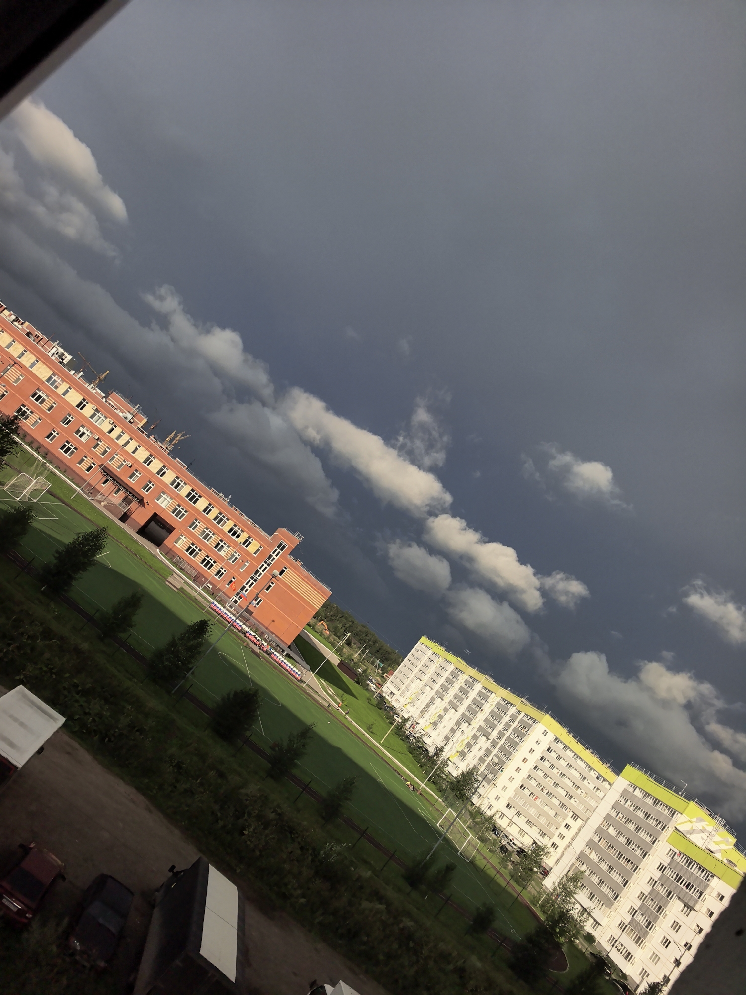 The image depicts a view of a residential area in the city of Froly, Russia, under a dramatic, cloudy sky. In the foreground, a dirt road is visible with several parked vehicles, including a red car, a dark sedan, and a large truck with a white rectangular structure on its back. Adjacent to the road is a grassy area with trees and the edge of a sports field, likely a soccer field, with white goalposts.
Dominating the middle ground are two multi-story buildings. The building on the left is a long, orange brick structure with numerous windows and several cranes visible on its roof, suggesting ongoing construction or maintenance. The building on the right is a modern, white apartment complex with green accents on its upper floors, also featuring many windows. The sports field stretches out between these buildings.
The background consists of a dark, stormy sky with patches of lighter clouds. The overall lighting suggests either pre-storm conditions or the aftermath of a storm, with strong contrasts between the dark sky and the illuminated buildings and field. The image is taken at an angle, giving a sense of looking down from a higher vantage point.