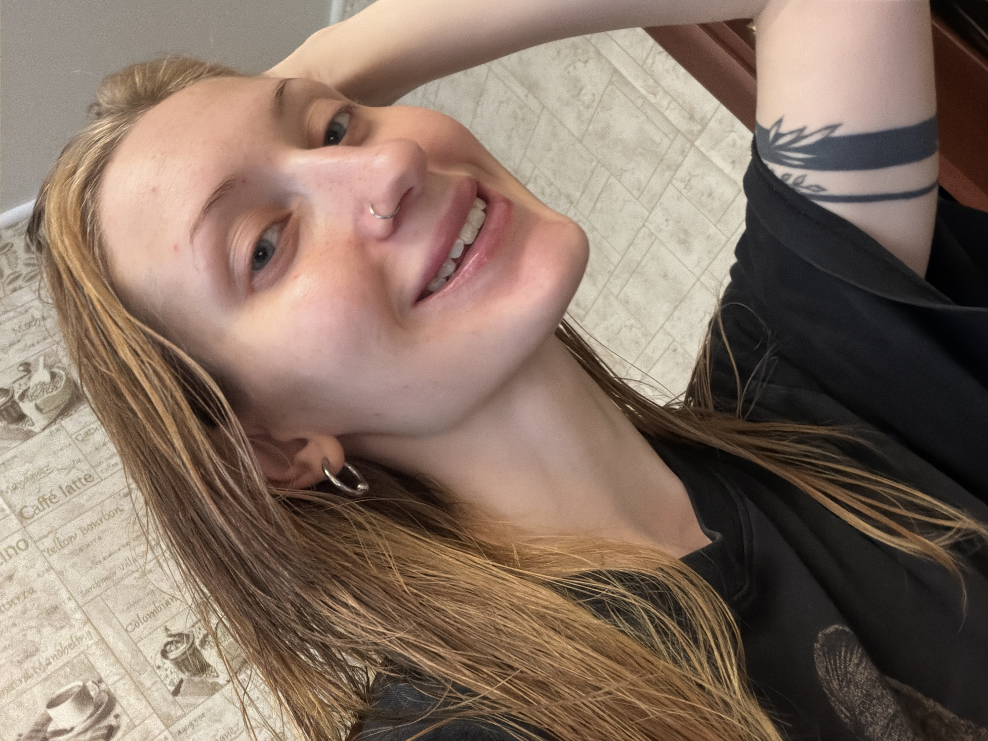 The image is a selfie of a young woman with long, blonde hair, taken from a slightly overhead, tilted angle. She is smiling widely, revealing her teeth, and looking towards the camera. She has a nose ring and a hoop earring in her visible ear. Her right arm is raised and bent, with her hand resting on the back of her head, partially obscuring her hair. She is wearing a black t-shirt, and a dark tattoo with bands and what appears to be a stylized plant or sun design is visible on her upper right arm.
The background appears to be indoors, with a tiled floor and what looks like a countertop or table covered with a patterned material. The pattern includes text and images related to coffee, with phrases like "Caffè latte," "Colombian," and "Maxwell House." The lighting suggests it could be daytime, but there are no specific indicators of the time of day or weather. The woman's expression conveys happiness and a relaxed demeanor. The overall setting seems casual and personal.