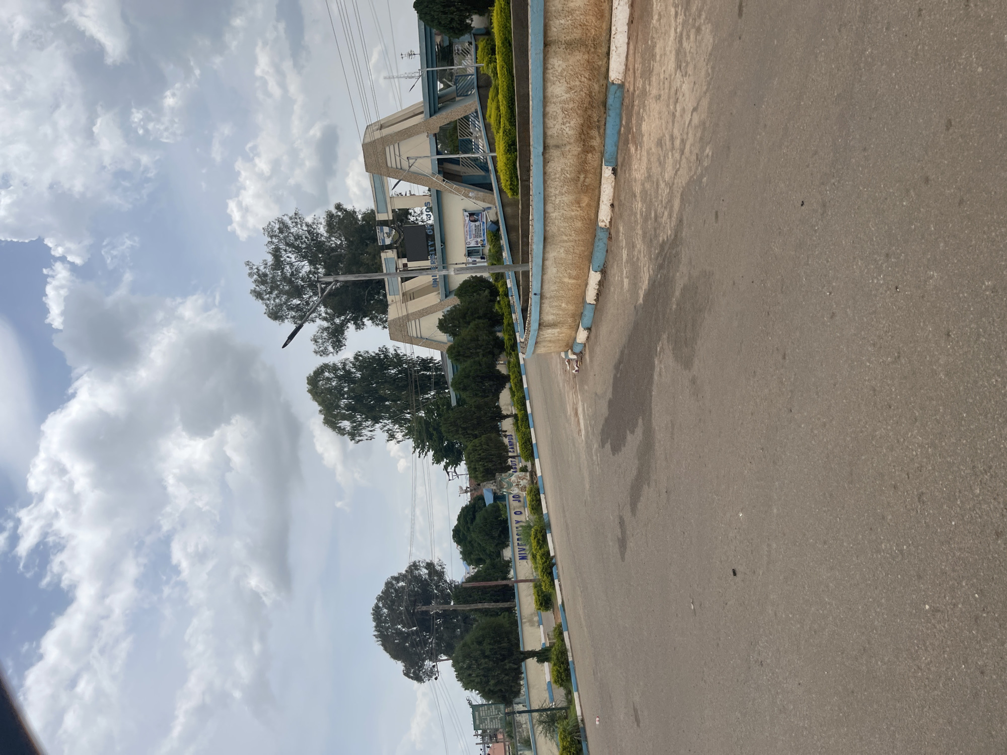 The image shows the entrance to the University of Jos, a public university located in Jos, Plateau State, Nigeria. The entrance features a large, modern archway with the university's name inscribed above the gateway. The university buildings are visible in the background, along with trees and lush green vegetation. The road in front of the university is paved and appears to be well-maintained. The sky is cloudy, suggesting a chance of rain. The scene is calm and orderly, with no people or vehicles visible in the immediate vicinity.