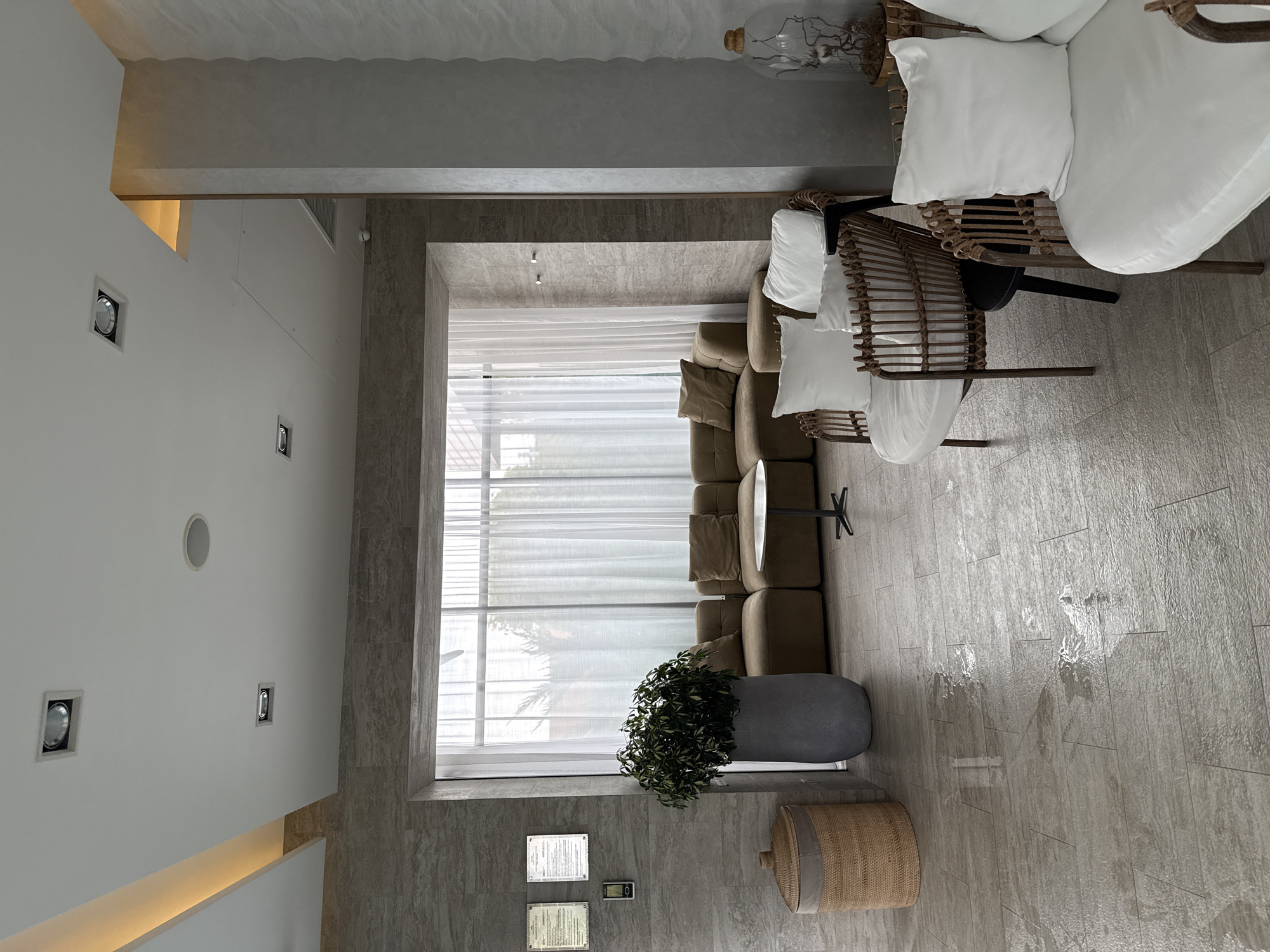 The image depicts an interior scene, likely a reception or lounge area. The flooring is made of large, light-colored rectangular tiles with a matte finish. There are several areas where the floor appears wet or reflective, suggesting recent mopping or spillage.
In the foreground, to the right, are two white upholstered chairs with woven rattan frames. In the middle ground, a large, plush modular sofa in a muted brown color is arranged in an L-shape, facing a large window covered by sheer white curtains. In front of the sofa, a small, round, dark-colored table with a central pedestal base is visible. To the left of the sofa, a large, dark gray planter holds a lush green plant. Further to the left, a woven basket with a fabric liner sits on the floor.
On the wall to the left, above the basket, are two framed documents or certificates mounted on a tiled surface with a stone-like texture. A small digital device is mounted below them. The ceiling features recessed lighting fixtures, including square and circular ones, and an integrated LED strip light illuminating an architectural element.
A tall, textured column with a rough, concrete-like finish stands on the right side of the image. On top of this column, a glass cloche encases decorative branches.
The overall aesthetic is modern and clean, with a focus on neutral tones and natural textures. The lighting is diffused, creating a soft ambiance. The scene appears to be unoccupied by people. The location is stated to be in Razumnoye, Russia.
