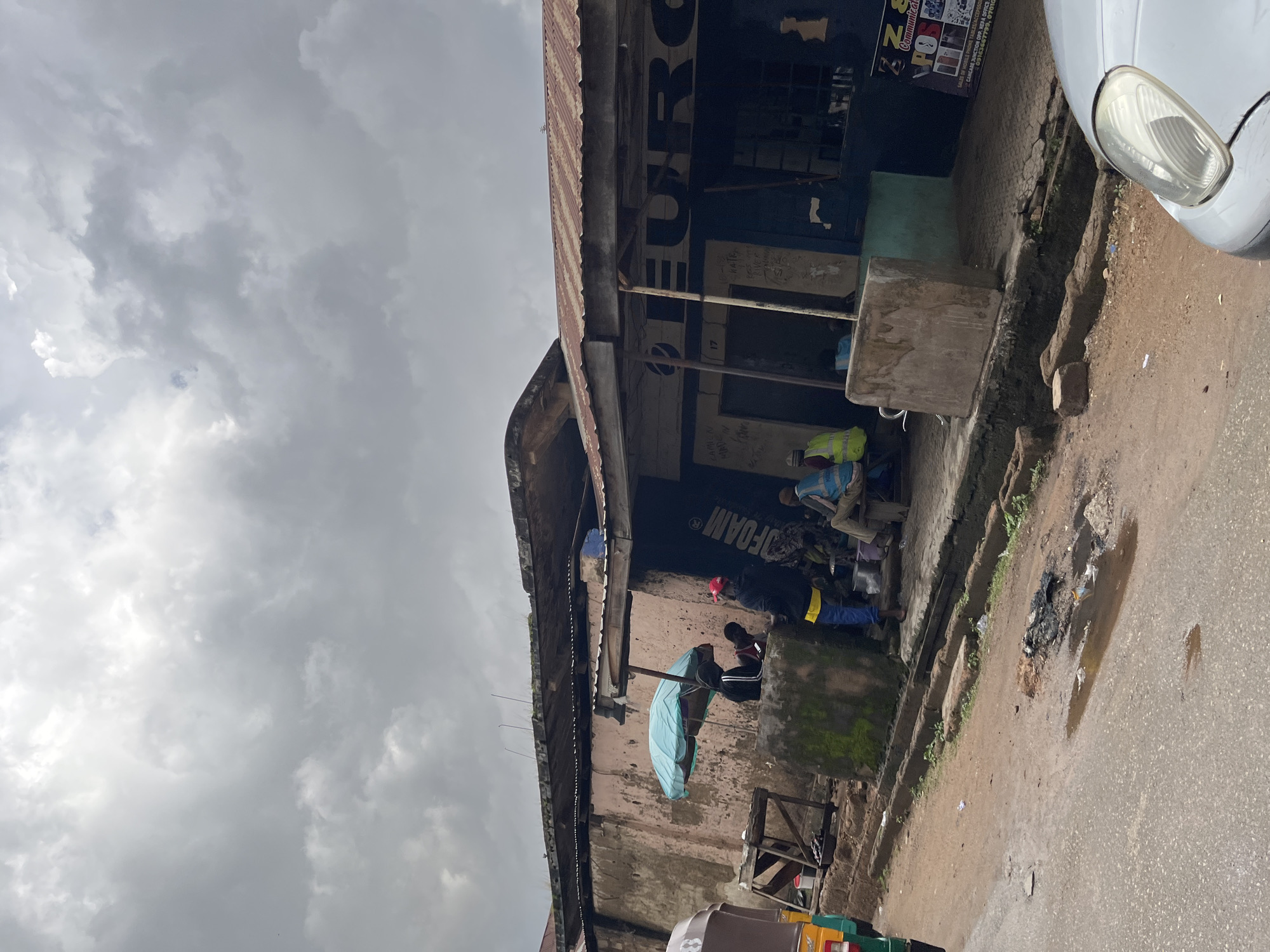 The image captures a street scene in Jos, Nigeria, under a cloudy sky, suggesting a potentially overcast or approaching rainy weather. The central focus is a weathered building with a corrugated metal roof, housing a shop. The shop's facade displays signage with the word "EURO" prominently featured and also "FOAM". Below this, further text, partially obscured and handwritten, seems to list contact information or services, including "CAMION MADE", "SALES OF MOBILE PHONE TABLES/ACCESSORIES", and phone numbers.
Several individuals are visible in the foreground and near the shop entrance. Some appear to be sitting or standing, engaged in activities that are not clearly discernible but suggest a daily life setting. One person is under a light blue umbrella, possibly seeking shelter from the elements or sun. Two individuals in the shop area are wearing bright yellow high-visibility vests, implying they might be workers or vendors.
The ground in front of the building is a mix of dirt and a paved surface, with a gutter running alongside the building. There are some puddles and debris on the ground, indicating the presence of water and perhaps recent rain or poor drainage. A portion of a white vehicle is visible on the right side of the frame, with its headlight illuminated. The overall impression is of a busy, everyday commercial area in a West African city.