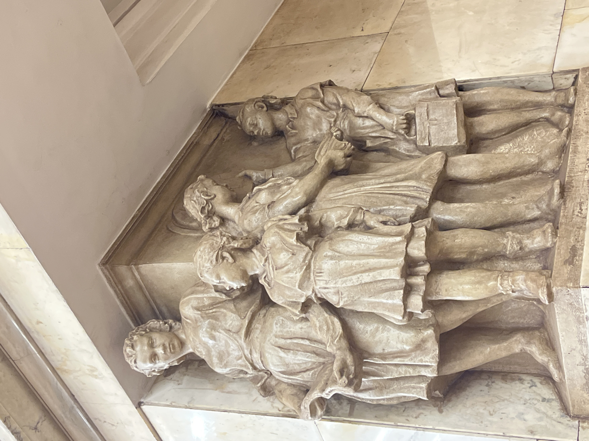 The image shows a low-relief sculpture of four figures, likely carved in stone or a similar material. They are positioned horizontally in a tiered arrangement, one on top of the other. The sculpture depicts what appears to be people in varying states of repose, possibly representing a scene of rest or mourning. The sculpture is integrated into a wall, likely an interior space, and seems to be a built-in part of the architectural structure. The location is identified as being in Saint Petersburg, Russia, but the specific location of the sculpture within the city is not visually apparent.