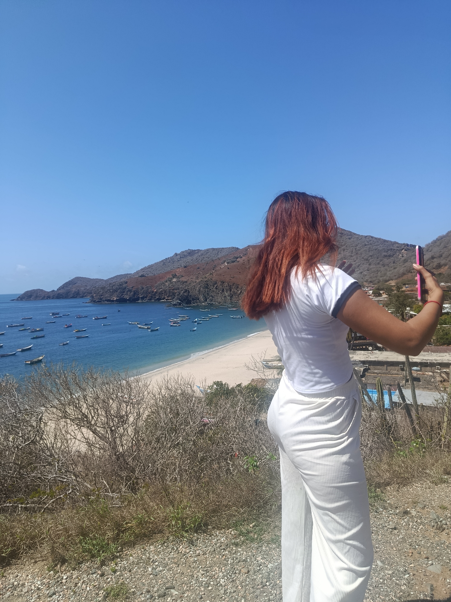 A woman with reddish-brown hair, seen from the back, stands on a rocky incline, taking a selfie with a pink smartphone. She is wearing a white t-shirt with a grey trim and white ribbed wide-leg pants. In the background, a scenic view unfolds with a crescent-shaped sandy beach, calm blue water dotted with numerous small boats, and rolling, arid hills that form a bay. Scattered buildings and cacti are visible on the hillside to the right, indicating a coastal settlement. The sky is clear and blue. The location is the city of Pedro González, Venezuela.