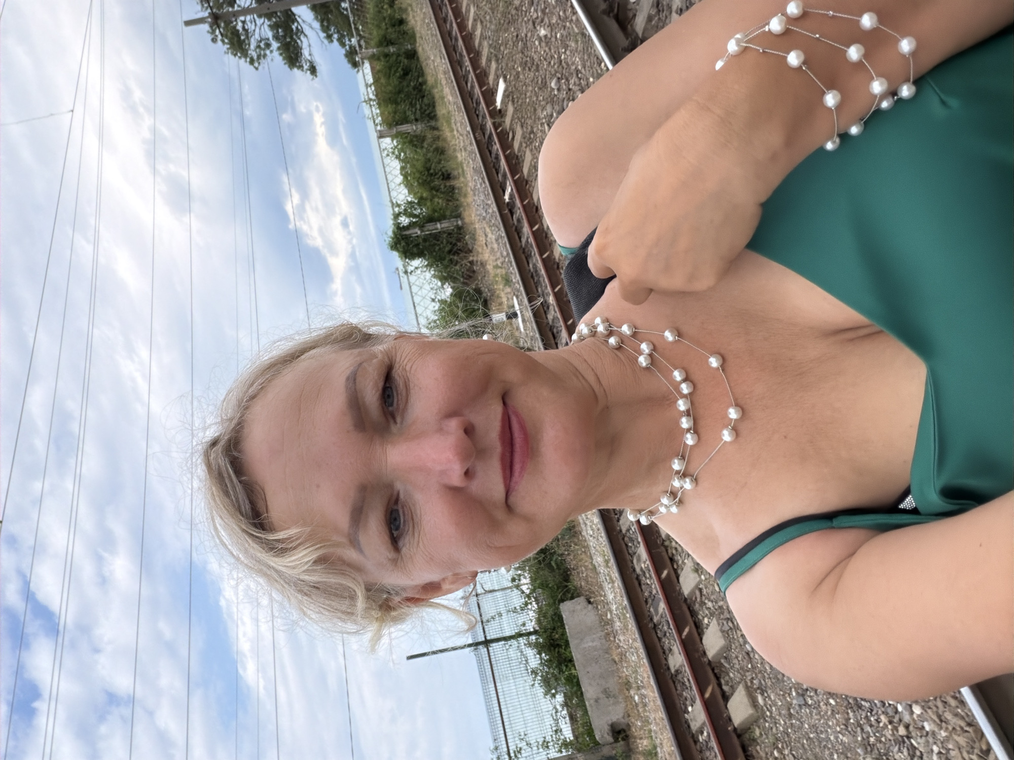 A woman with blonde hair and fair skin, wearing a green dress, a pearl necklace, and a pearl bracelet, takes a selfie. She is standing on a gravel surface next to a set of train tracks. In the background, a metal fence runs along a body of water, beyond which is a cloudy sky with visible power lines. The location is Shepsi, Russia.