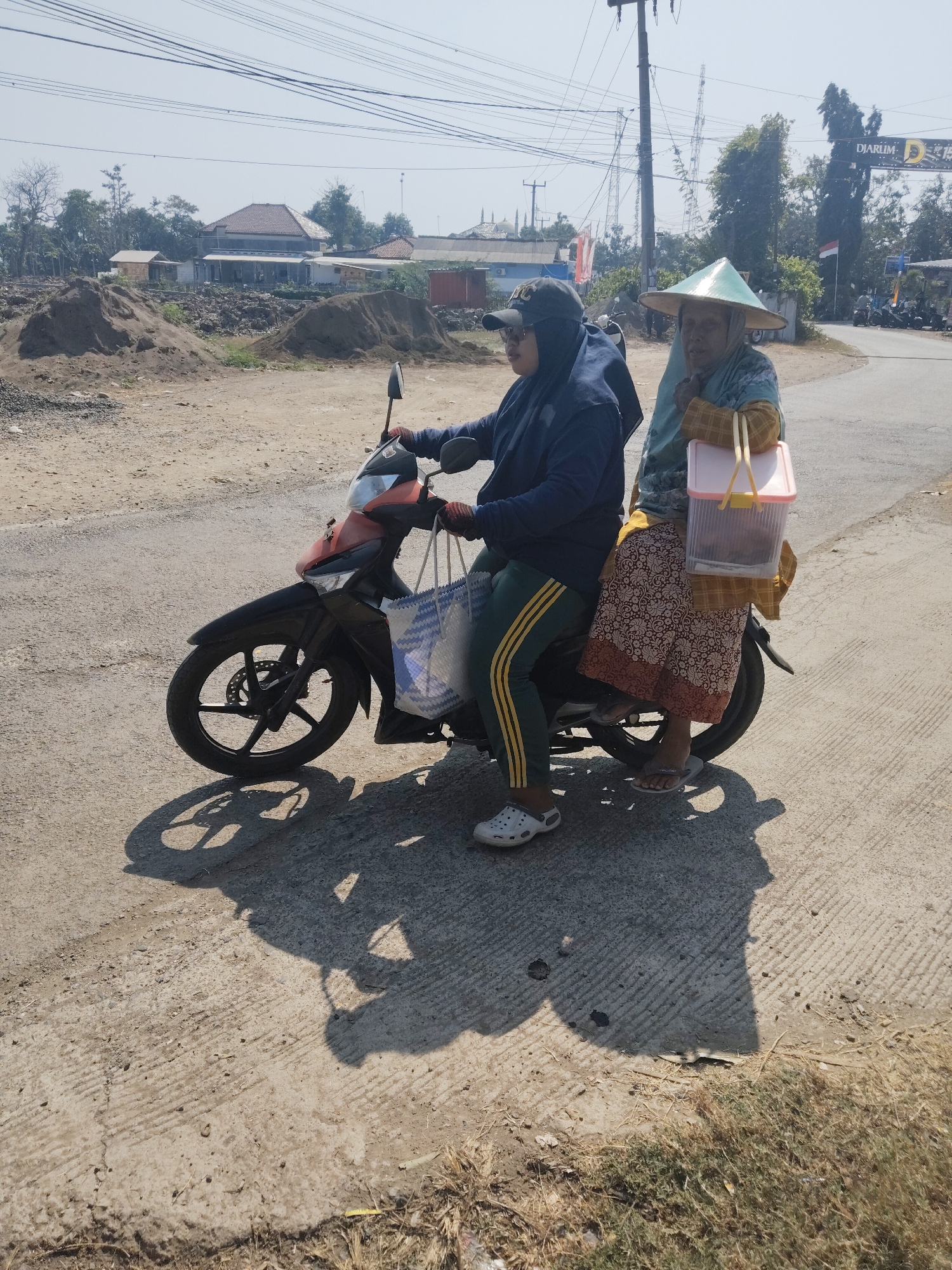 Two individuals, one driving and one as a passenger, are seated on a red and black motorcycle on a paved road. The driver wears a dark blue hooded jacket, green pants with yellow stripes, and white Crocs. The passenger, wearing a traditional conical hat and a patterned skirt, holds a pink plastic container with yellow handles. The background shows a rural or semi-urban setting with construction materials, houses, trees, and power lines under a clear sky. A sign for "DJARUM" is visible in the distance. The scene appears to be in the city of Subang, Indonesia.