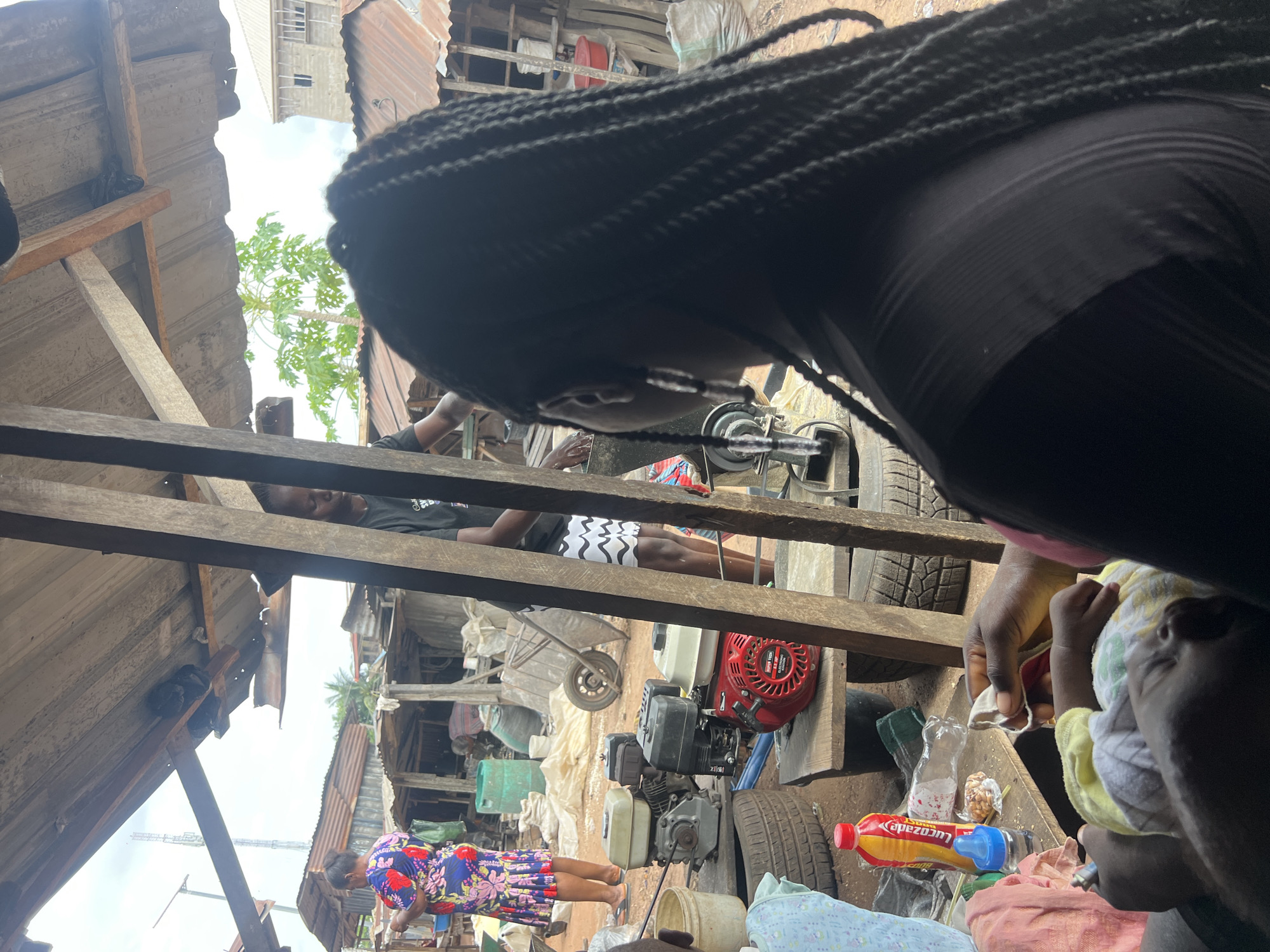 The image shows an outdoor scene in a cluttered urban environment, possibly a marketplace or a low-income residential area of Unknown, Unknown.
Several people are visible: A woman in a blue floral dress is walking near the bottom of the frame, away from the camera. Another individual is lying on wooden beams in the center of the image. A baby and a hand holding the baby are to the right. A woman with black braided hair is partially obstructing the upper portion of the view.
The environment is characterized by structures made of corrugated metal and wood, with evidence of makeshift construction and general disrepair. Visible objects include car tires, a gas-powered engine, bottles, buckets, and other household items cluttered in the foreground. Wooden beams and makeshift roofing materials define the immediate surroundings. Overall, the scene depicts a densely populated and somewhat impoverished urban landscape.