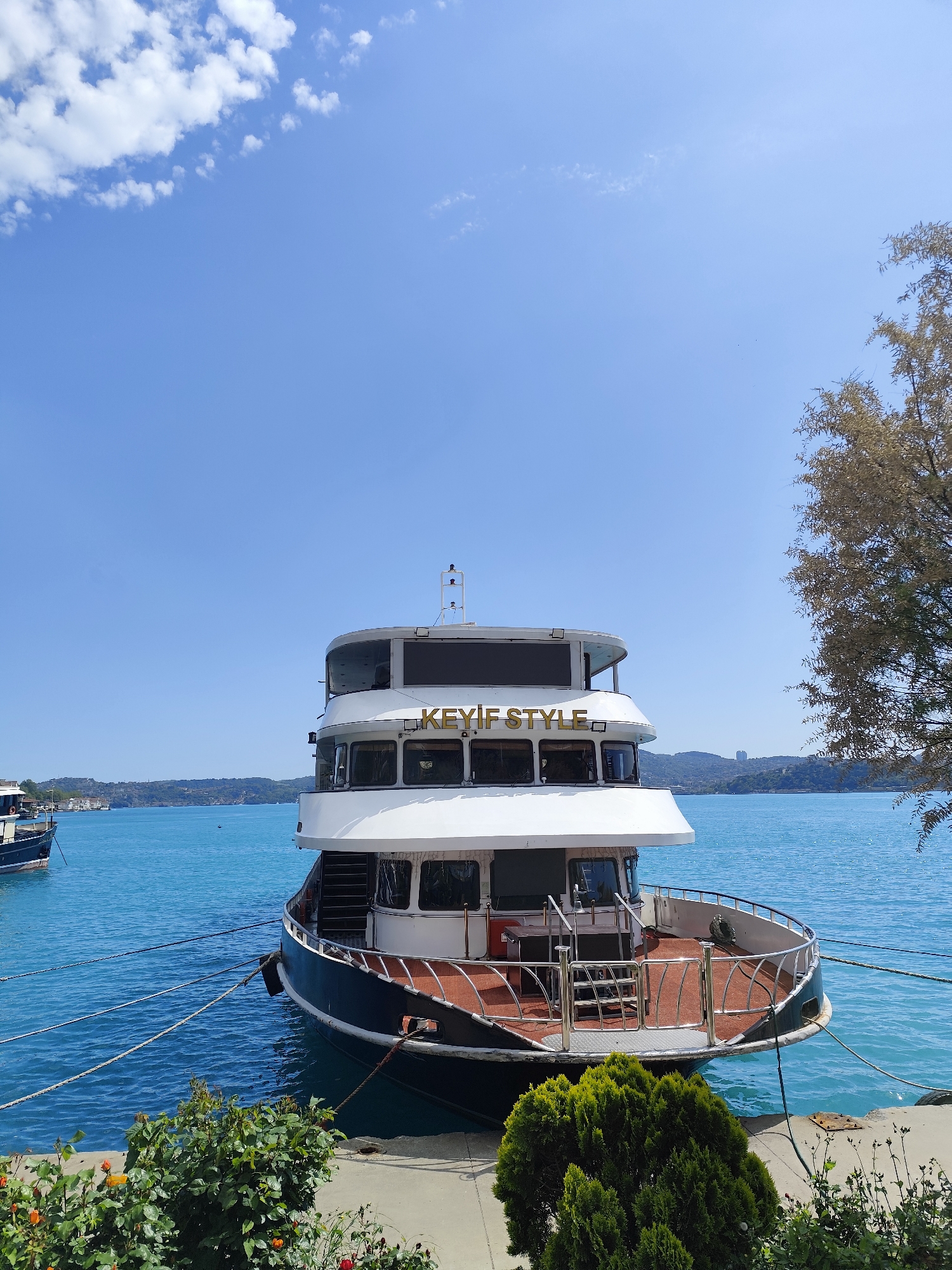 A three-story boat named "KEYIF STYLE" is docked at a pier in Sarıyer, Turkey. The boat has a white upper structure and a dark lower hull. It is secured with ropes to the concrete pier. The deck is brown and has a chrome railing. A smaller boat is visible in the distance. The water is a bright turquoise color. A tree with sparse leaves is on the right. The pier is lined with small plants. The sky is blue with scattered white clouds. Hilly land is visible across the water in the background.