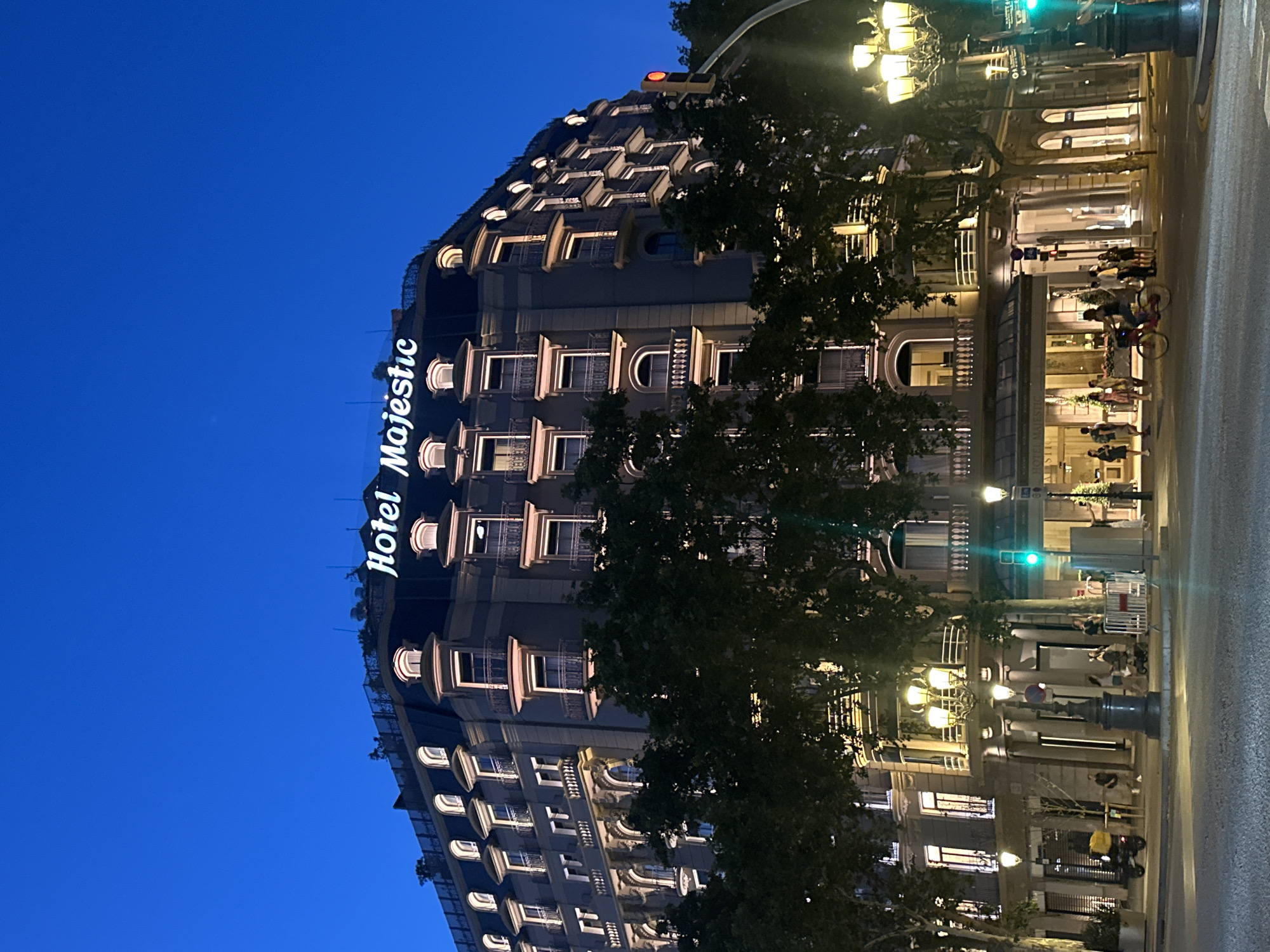 The image depicts the exterior of the Hotel Majestic in Barcelona, Spain, at dusk. The ornate architecture of the hotel is illuminated, with warm light emanating from its many windows. The hotel's name, "Hotel Majestic," is prominently displayed in glowing white letters on its facade.
In front of the hotel, a street scene unfolds. Several people are visible, some standing and others walking. A few individuals are riding bicycles, suggesting a leisurely activity or commute. Streetlights cast a warm glow on the pavement, contributing to the evening ambiance. A traffic light is visible, showing a green signal.
The time of day is clearly evening, given the deep blue of the sky and the artificial lighting in use. The weather appears to be clear. The presence of trees with dense foliage adds to the urban landscape. The scene conveys a sense of a vibrant city street at the end of the day, with people engaging in various activities. The overall mood is calm and inviting, with the illuminated hotel serving as a focal point.