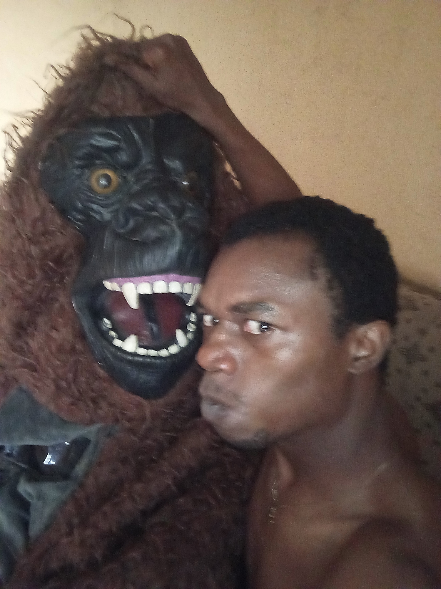 A portrait-style image features a man and a gorilla mask/costume in close proximity. On the right, a dark-skinned man with short, dark hair is visible from the chest up, facing partially left with an intense or serious expression. He appears to be shirtless, wearing a thin, golden chain around his neck. His face is angled, with his gaze directed towards his left (viewer's right).
To the left of the man is a prominent gorilla mask and what appears to be part of a furry brown costume. The mask is dark, almost black, with large, piercing yellow eyes. Its mouth is wide open, revealing bright white teeth, including exaggerated fangs, and a pinkish interior. A dark-skinned human hand rests on top of the brown, shaggy fur of the gorilla costume, behind the mask's head. The positioning suggests the gorilla mask is either being worn by someone or held up next to the man, or the man himself is wearing the costume.
The background is a plain, light beige or tan wall, with a hint of a patterned fabric (possibly a sofa or cushion) visible behind the man's right shoulder. The lighting is somewhat dim, giving the scene an intimate or slightly moody feel. There is no discernible text in the image.