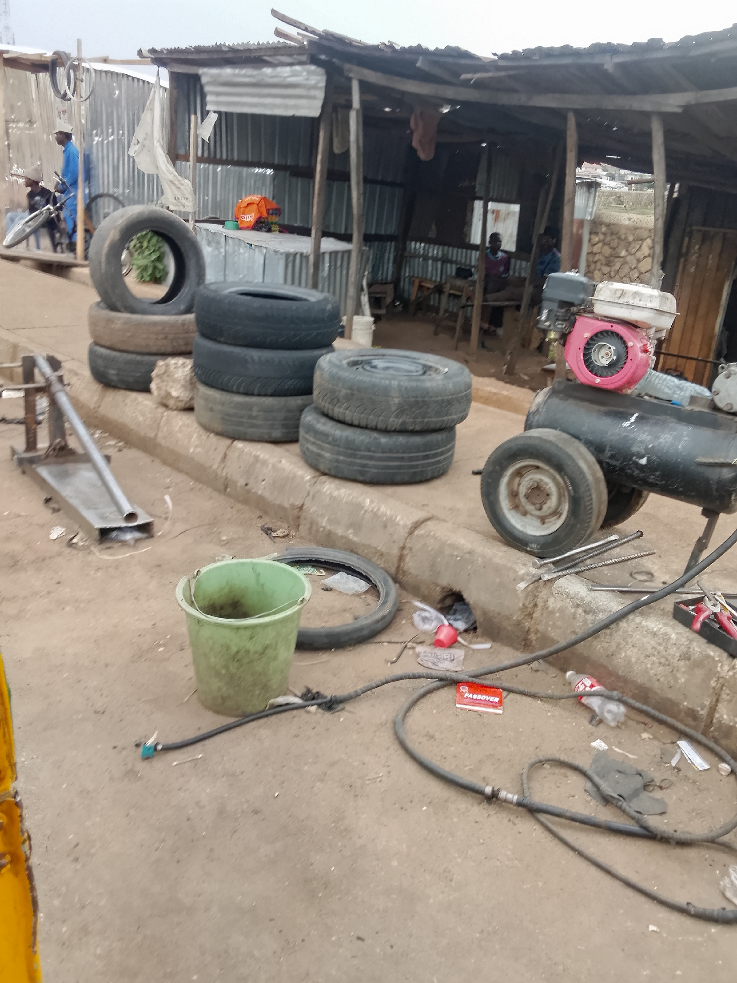 The image depicts a roadside tire repair shop in Bukuru, Nigeria. The shop is situated on a concrete street corner, with a makeshift shelter constructed from corrugated metal and wooden supports. Stacks of used tires are piled along the edge of the street, marking the shop's location. A black air compressor with a pink engine sits nearby, along with various tools and a green bucket. Litter is scattered on the ground, including plastic bottles and wrappers. In the background, a man in a blue shirt appears to be working on a bicycle. Other people are seated inside the shelter, possibly waiting for service.