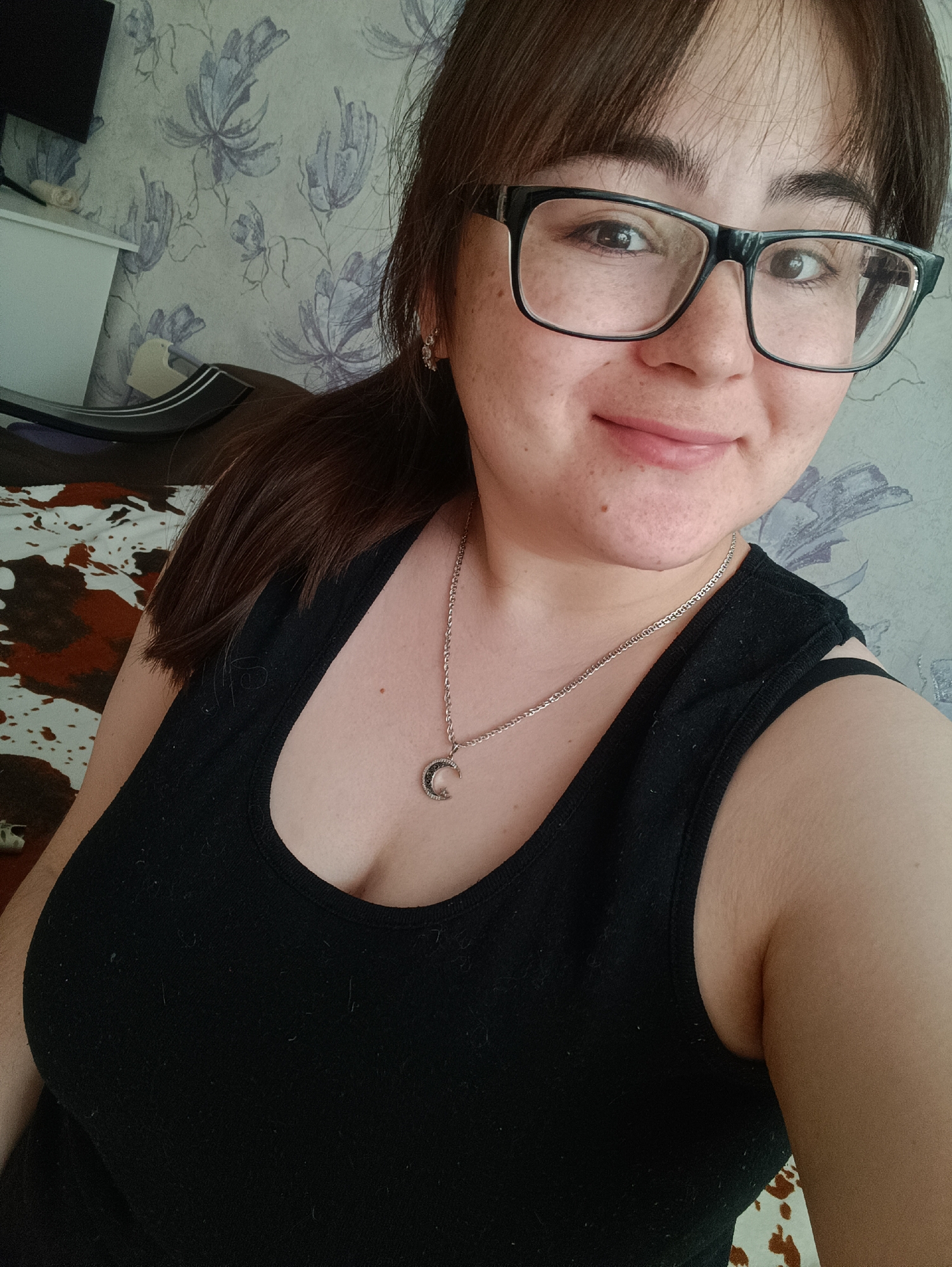 A young woman in Noviy, Russia, is depicted in a close-up, chest-up shot, likely a selfie, in an indoor setting. She has fair skin with visible freckles and a gentle, closed-mouth smile. Her dark brown hair is styled with bangs across her forehead, and some of it is pulled back, with a portion of a ponytail visible on the left side. She is wearing black, rectangular-framed glasses and a black tank top. Around her neck is a silver-colored chain necklace with a crescent moon pendant, and a small earring is visible in her right ear.
The background features a light-colored wall with a repeating pattern of stylized blue-grey floral or botanical designs. To the left, a portion of white furniture, possibly a cabinet, is visible, with a dark electronic device on top. A brown and white patterned fabric, suggestive of a bedspread or blanket, is partially seen behind her. The lighting appears natural, indicating daytime. No text is visible in the image.