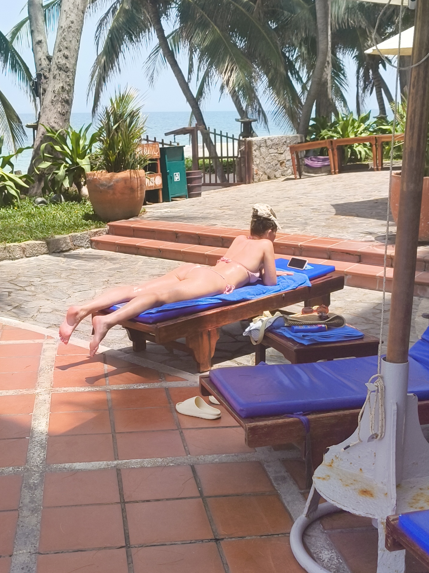 In Phu Thuy Ward, Vietnam, a woman wearing a pink bikini lies on a wooden lounge chair on a tiled patio, positioned near a swimming pool and a view of palm trees and the ocean. The woman is on her stomach with her head turned to the side, looking at a tablet device. A blue towel is beneath her. Next to her on a lower table, a straw hat and sunglasses are visible. In the background, the ocean is visible beyond a fence and several palm trees. The patio is made of red tiles with grey grout.