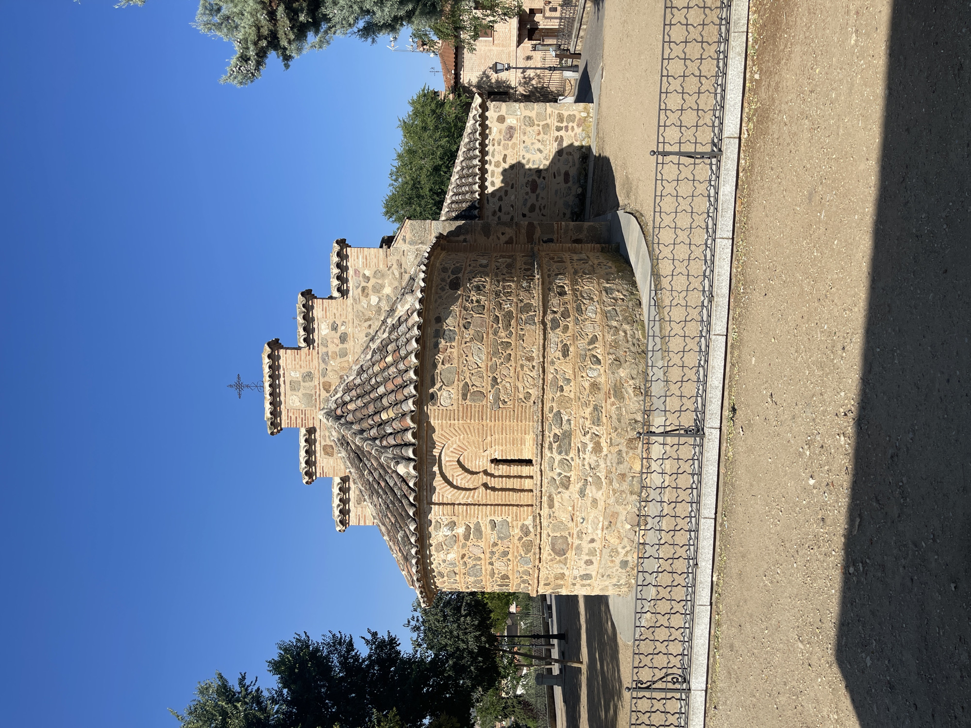 The image depicts the exterior of the Church of Santa María de Guadamur, located in the city of Guadamur, Spain. The church, constructed with rough-hewn stone and brickwork, features a rounded apse topped with a tiled roof. Above the apse, a stepped structure rises, adorned with clay tiles and culminating in a cross. A distinct arched window with brick detailing is visible on the apse's facade. The building is surrounded by a paved area and enclosed by an ornate black metal fence. Trees, including an evergreen, are visible to the left and right of the structure, with a clear blue sky overhead. In the background, more buildings and vegetation can be observed.
