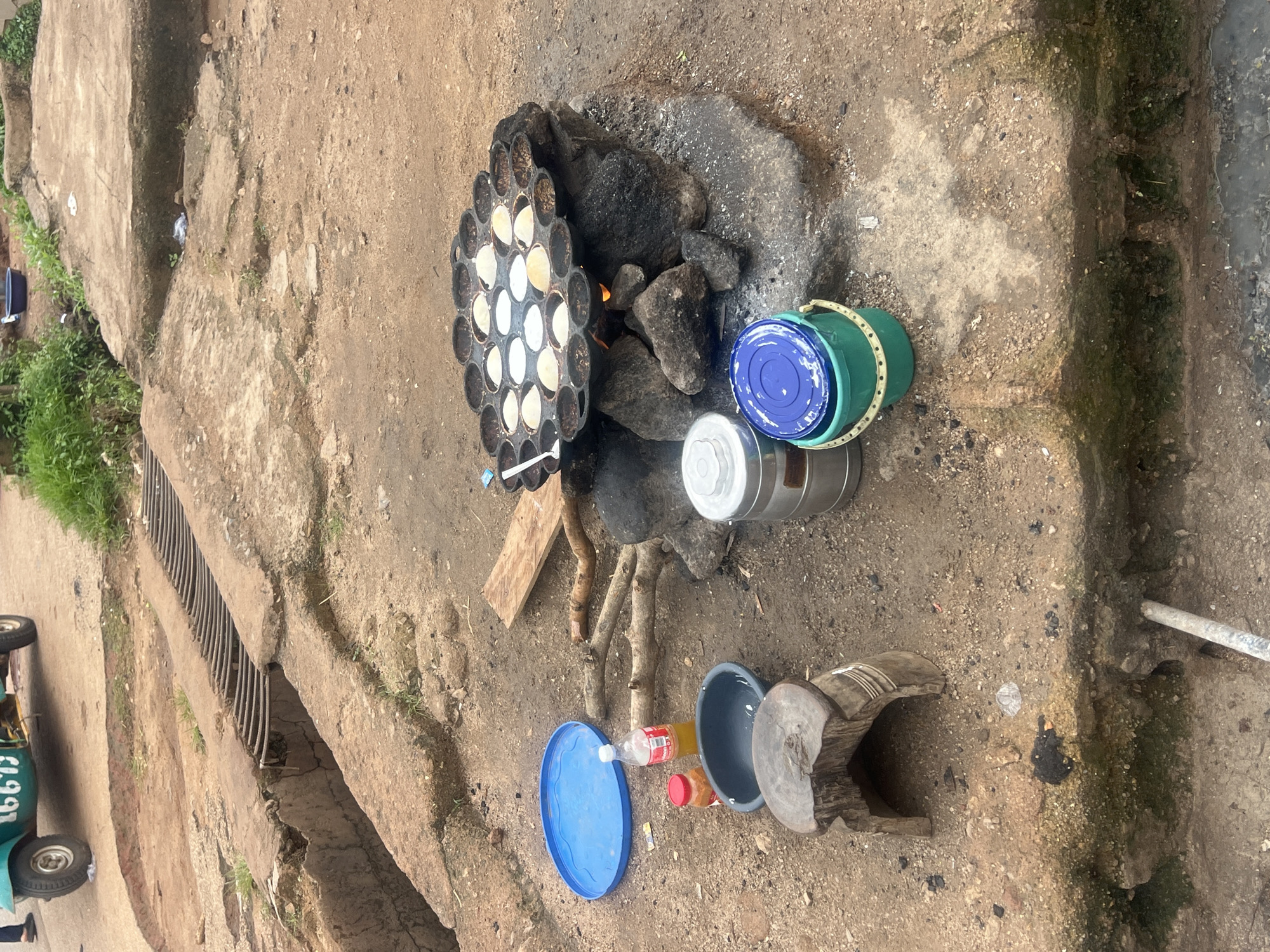 The image depicts an outdoor cooking scene, likely in Jos, Nigeria, characterized by a rustic, earthy environment. In the foreground, a traditional cooking setup features a cast-iron pan with numerous small indentations, filled with what appears to be a batter for cooking pancakes or a similar item. This pan is placed over a makeshift stove constructed from rocks and burning embers, producing visible heat and smoke.
Beside the cooking area, several objects are arranged on the dirt ground. These include a bright blue plastic plate, a bottle of beverage, a small jar with a red lid, and a dark grey bowl. A small, crudely carved wooden stool is positioned nearby. Further to the right, two metallic containers, one taller and silver, and a shorter, wider green bucket topped with a blue lid, are present.
The background reveals elements of the urban environment, with a drainage grate and a partially visible green and white vehicle with registration number "16671" suggesting proximity to a road. The ground is uneven, consisting of dirt, sand, and some scattered debris, with patches of green vegetation and moss indicating moisture. The overall impression is one of informal, everyday life, with food preparation taking place in an open, utilitarian space.