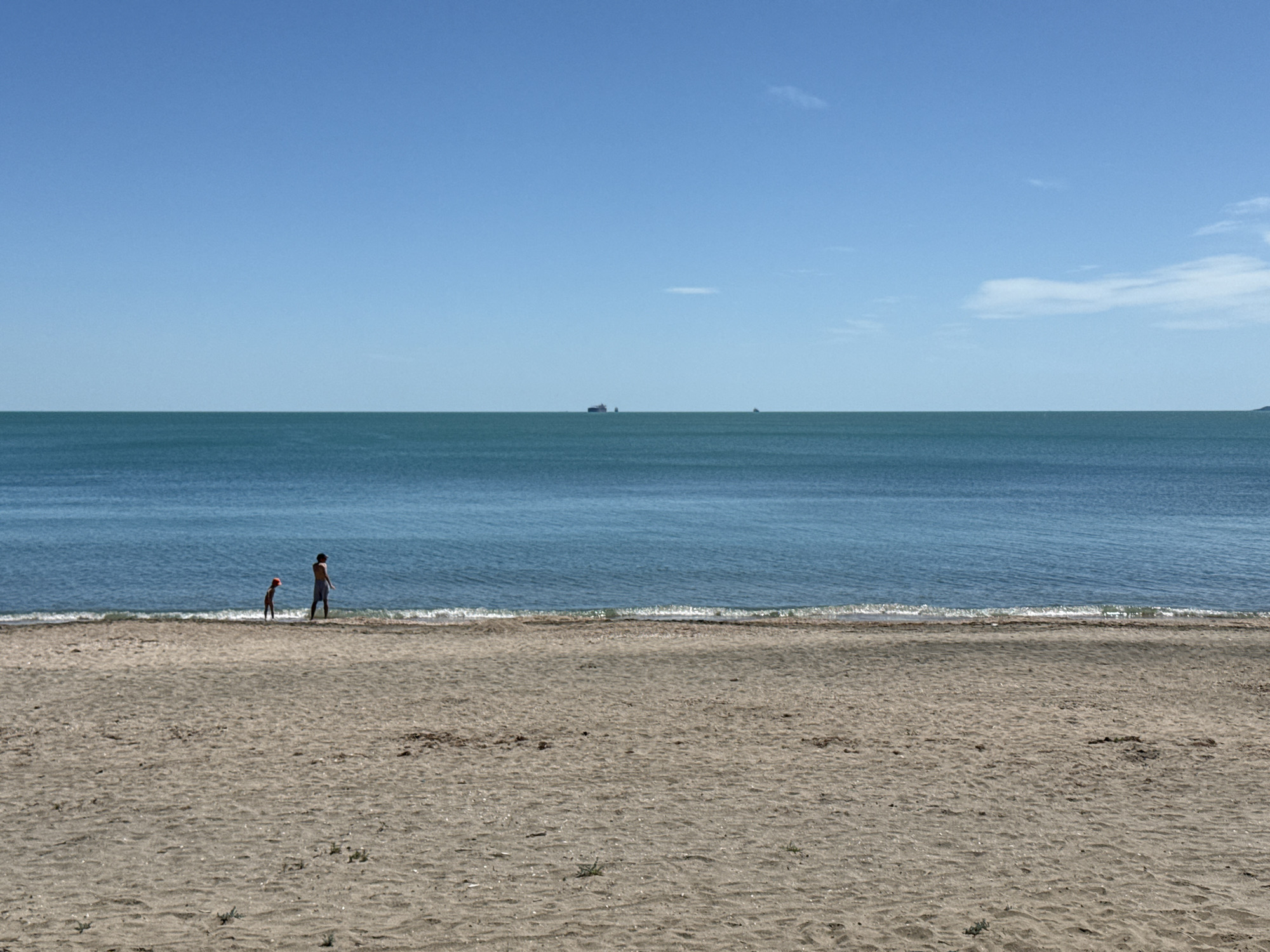 The scene is a wide-angle shot of a beach in Burgas, Bulgaria, under a clear blue sky. The foreground consists of a sandy beach with visible footprints and sparse vegetation. The middle ground shows the sea with gentle waves lapping at the shoreline. Two people, a child and an adult, are standing at the water's edge. In the background, the horizon features the sea meeting the sky, with a few distant ships and a landmass visible. The general atmosphere is calm and bright.