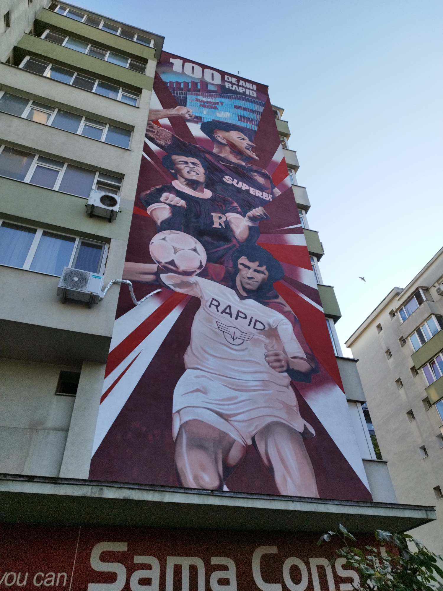 This is a photograph of a mural painted on the side of a multi-story building in Bucharest, Romania. The mural depicts three stylized figures of soccer players, with one figure in the foreground holding a soccer ball on his knee. The mural also includes text that reads "100 DE ANI RAPID," likely celebrating a 100-year anniversary of a soccer club. The building itself is light-colored with multiple windows and air conditioning units visible. The sky is clear and blue. The mural covers a significant portion of the building's facade.