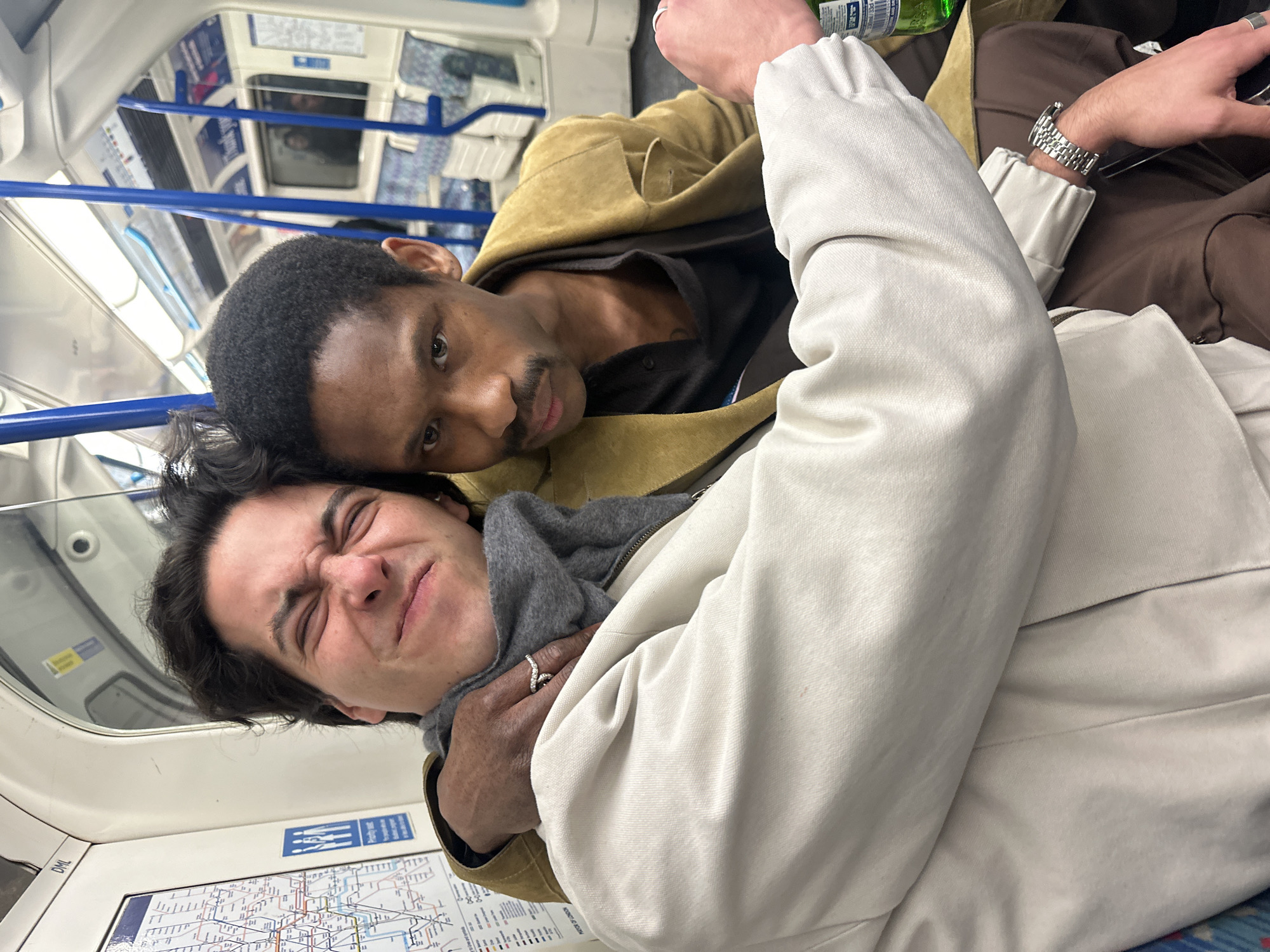 Two males are depicted inside a London Underground train carriage in London, United Kingdom. The train interior features a visible London Tube map, a "Priority seat" sign, blue vertical support poles, and patterned blue and white seating. The male in the foreground, with dark hair and light skin, wears a light-colored jacket and a grey scarf. He makes a humorous, contorted facial expression with eyes squeezed shut. A silver-toned ring with stones is on his left ring finger, and a silver-toned watch is on his left wrist. He holds a green bottle in his right hand. The second male, with dark skin, short dark hair, and a mustache, is positioned behind the first. He wears a mustard yellow jacket over a dark shirt and looks directly at the camera with a neutral expression. His right arm is placed around the shoulder of the male in the foreground.