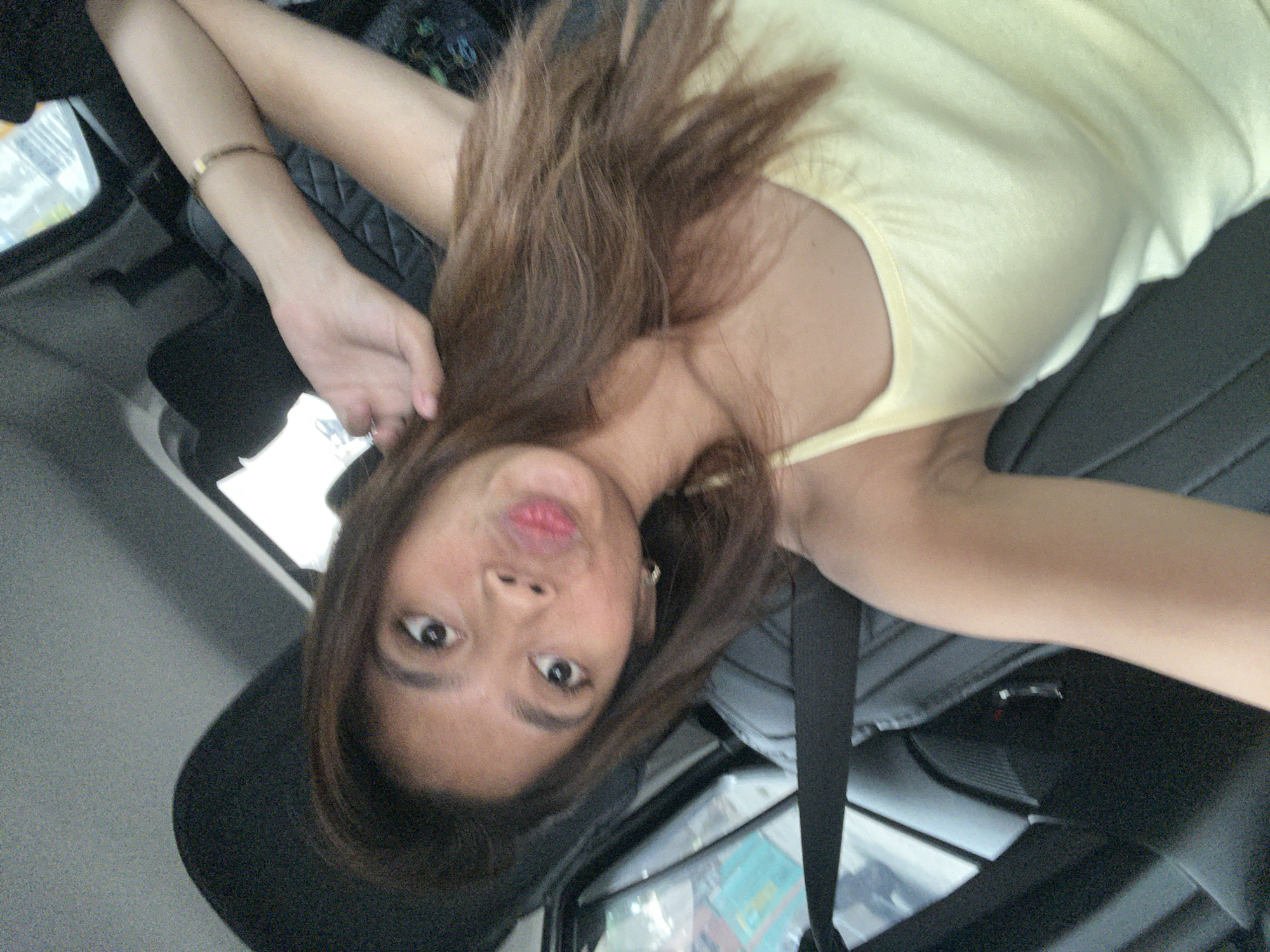 A woman is taking a selfie in a car. She has long, brown hair and is wearing a yellow tank top. She is making a kissing face. The car appears to be in motion, as indicated by the blurred background. The image was taken in Davao City, Philippines.