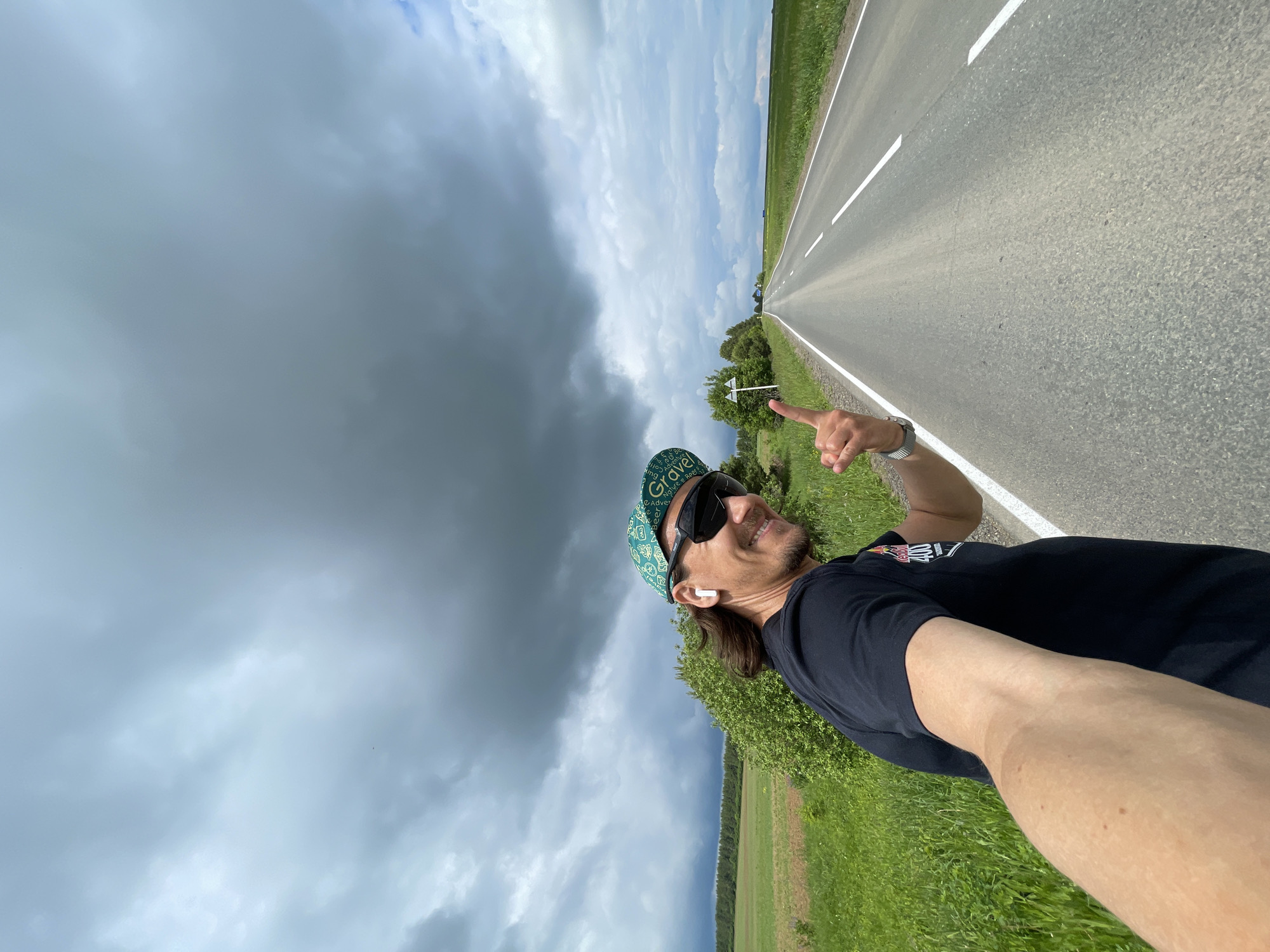 The image, oriented in a portrait style, features a man taking a selfie outdoors by a long, empty road. The man is positioned on the left side of the frame, smiling and looking towards the right. He has long hair, a beard, and is wearing sunglasses, a black t-shirt, and a green-and-white patterned baseball cap. White wireless earbuds are visible in his ears. His right arm is extended, with his index finger pointing into the distance, following the line of the road. A watch is visible on his left wrist.
The setting includes an asphalt road with white markings that extends from the bottom right corner towards the top right, disappearing into the horizon. Green grassy fields line both sides of the road. In the mid-ground, on the right, there are some trees and bushes, with a small white road sign partially visible among them.
The sky is a notable element, presenting a dramatic contrast. Large, dark, and heavy clouds dominate the left side of the frame and extend overhead, suggesting an approaching storm or overcast conditions. However, towards the top right, beyond the distant horizon, the sky is clearer with visible patches of blue and lighter clouds. The scene depicts a rural or open landscape during daytime. The man's cap has the word "Gravel" along with other text. His cheerful expression, coupled with the pointing gesture, suggests a sense of journey or adventure.