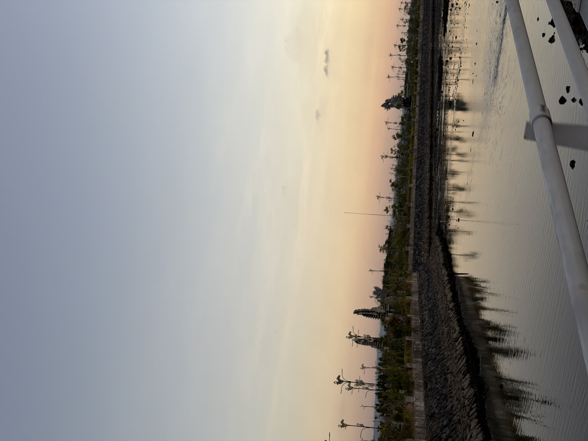 The image depicts a calm body of water reflecting the soft hues of a twilight sky. A stone embankment lines the water's edge, topped with a low fence and then a row of stylized trees or light fixtures resembling birds. In the background, several ornate statues, possibly traditional Indonesian figures, are visible amidst lush greenery. The scene suggests a public park or waterfront area in South Denpasar, Indonesia, at the end of the day.